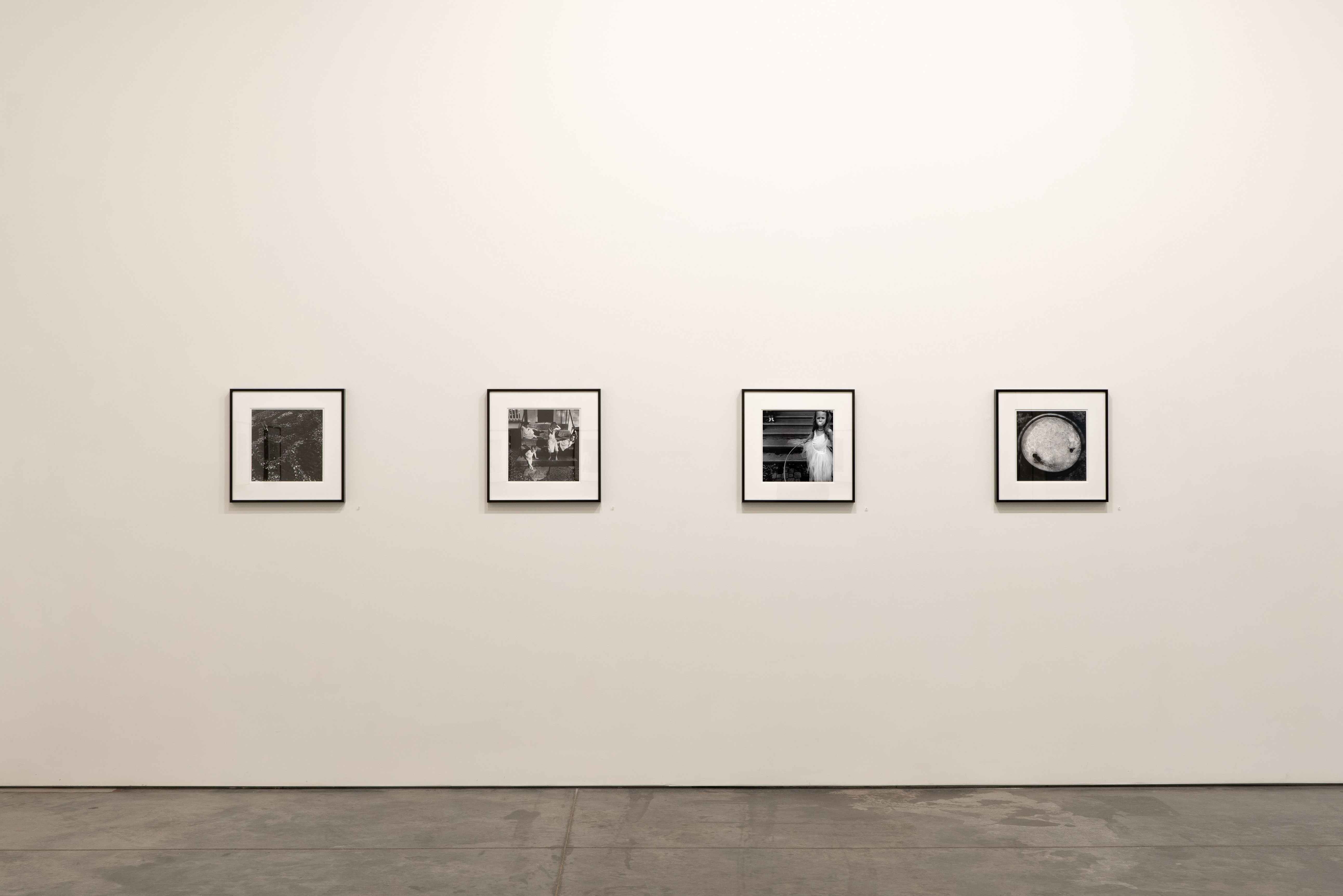 Four black and white framed photographs hang evenly spaced on a beige gallery wall at eye level. Each photograph, presented in a square format with white matting and black frames, depicts intimate domestic or personal scenes. The images appear to be from the early 2000s and display a documentary-style aesthetic.