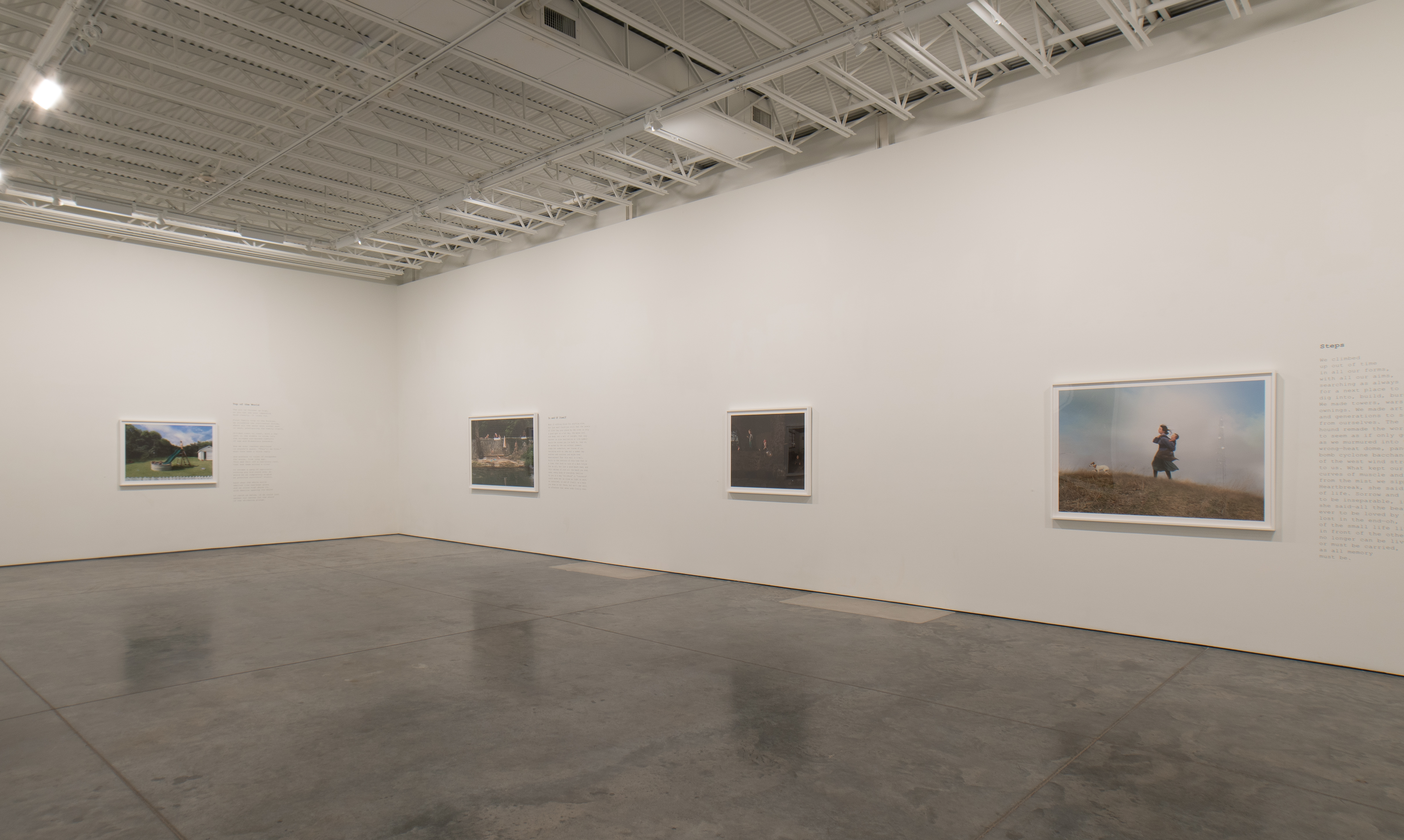A sparse gallery space with polished concrete flooring displays four framed photographs spaced across white walls beneath an industrial ceiling with exposed beams and skylights. Three smaller images hang on the left and center walls, while a larger color photograph of a figure on a hillside dominates the right wall, each accompanied by descriptive text plaques.