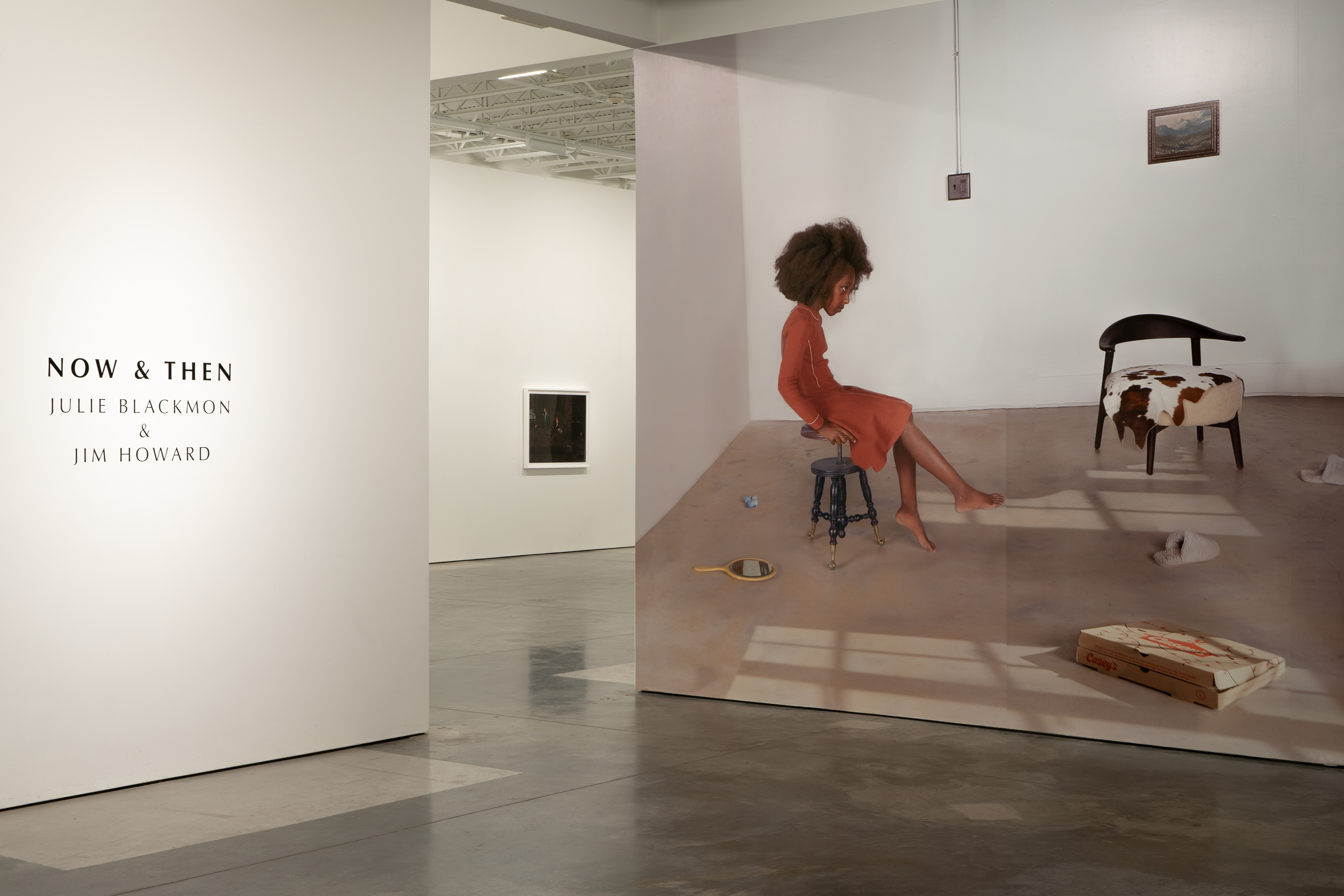 A gallery installation features a large-scale photograph depicting a child in a red dress sitting on a stool in a sparse interior space with various objects scattered on the floor, including books and a chair with patterned upholstery. White gallery walls display the exhibition title and artist credits on the left side.