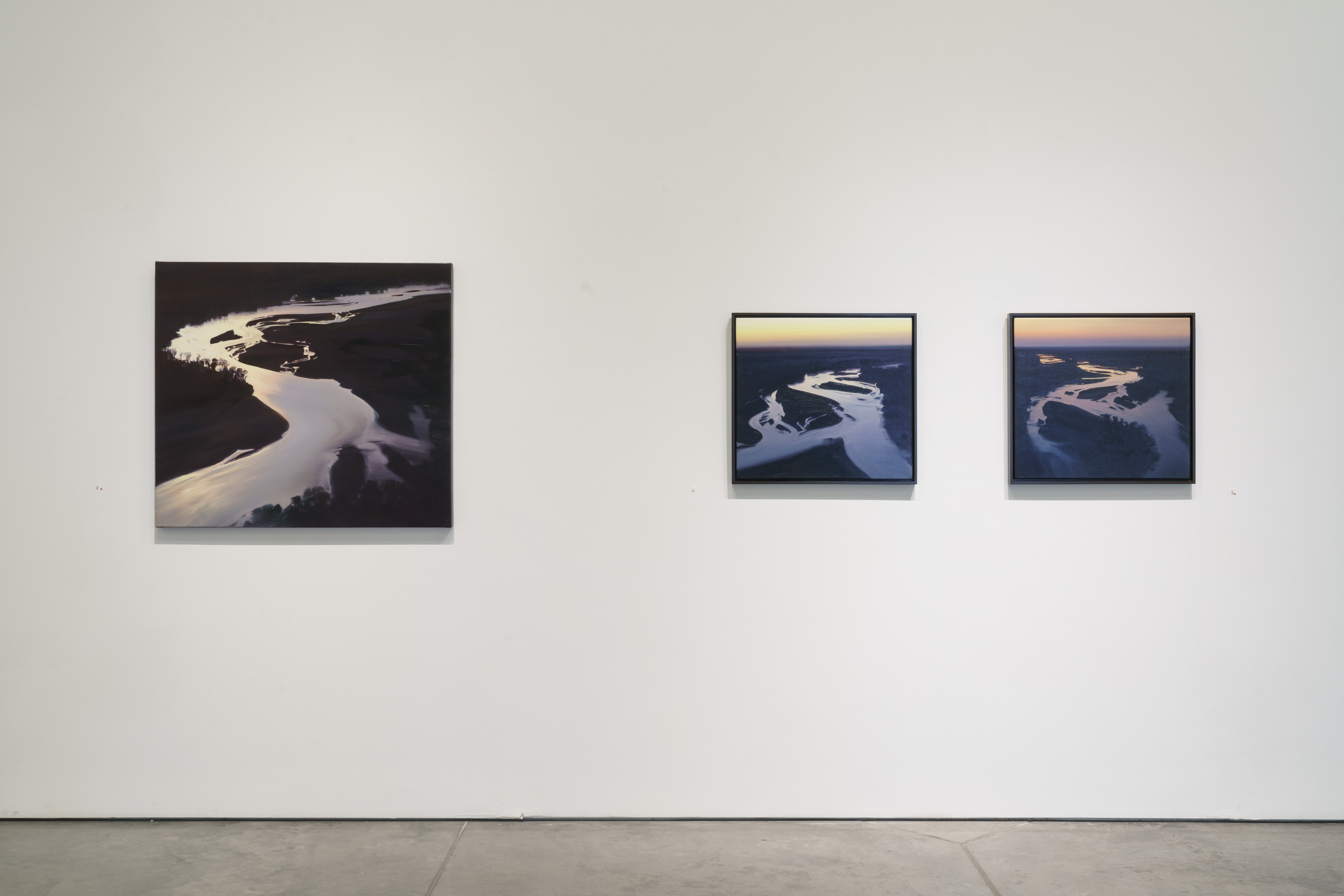 Three oil paintings depicting aerial views of a winding river with sandbars and surrounding landscape. The largest work on the left features dramatic contrast between luminous white water reflections and deep blacks of the land, rendered with visible brushstrokes. The two smaller framed paintings on the right show similar river perspectives in twilight tones of blue and purple with golden light at the horizon.