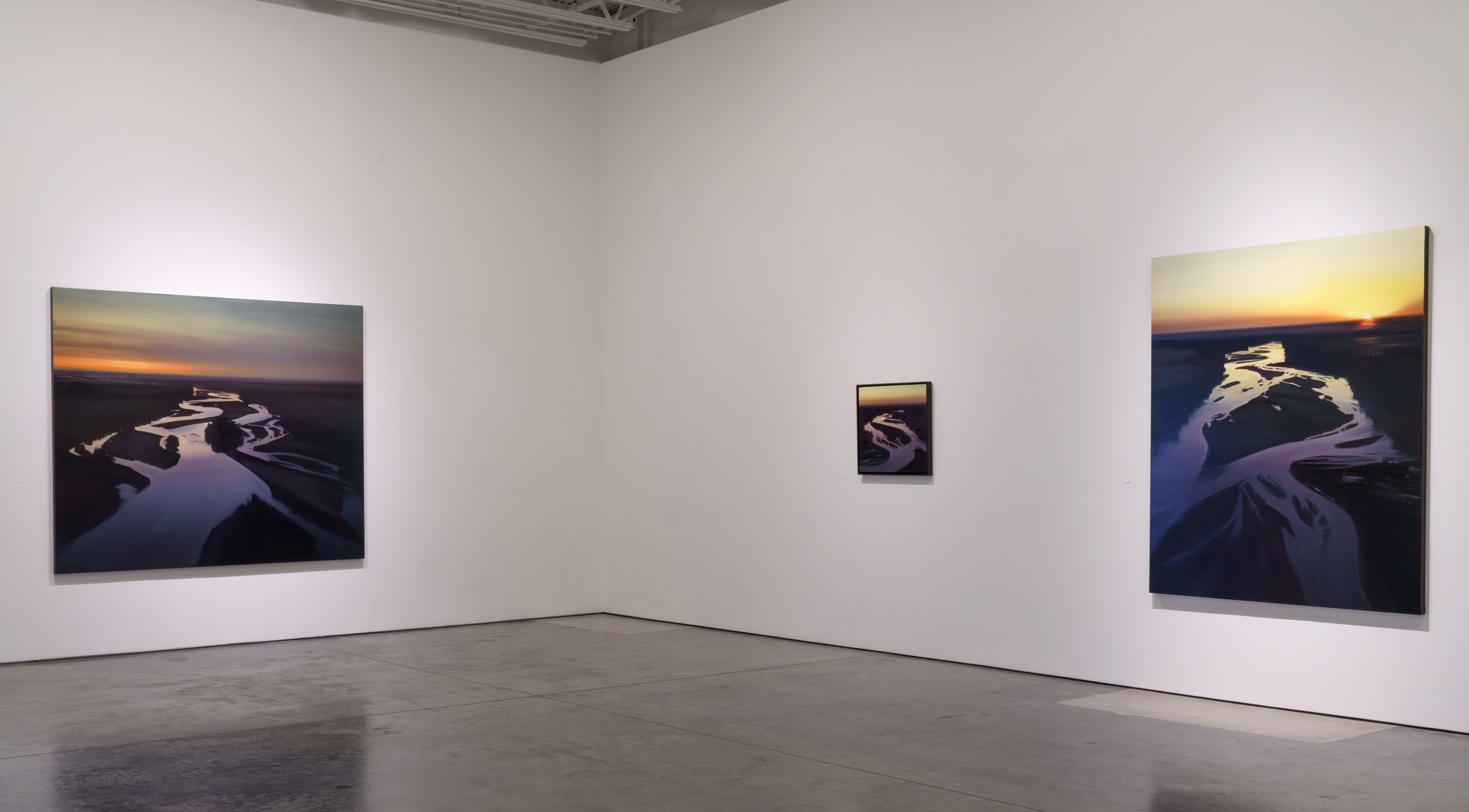 Three oil paintings displayed on white gallery walls depicting aerial views of a winding river at sunset and dusk. The compositions feature luminous whites and warm golds reflecting off water contrasted against deep, richly textured blacks representing sandbars and land, with brushwork that creates shimmer and optical depth across the canvases.