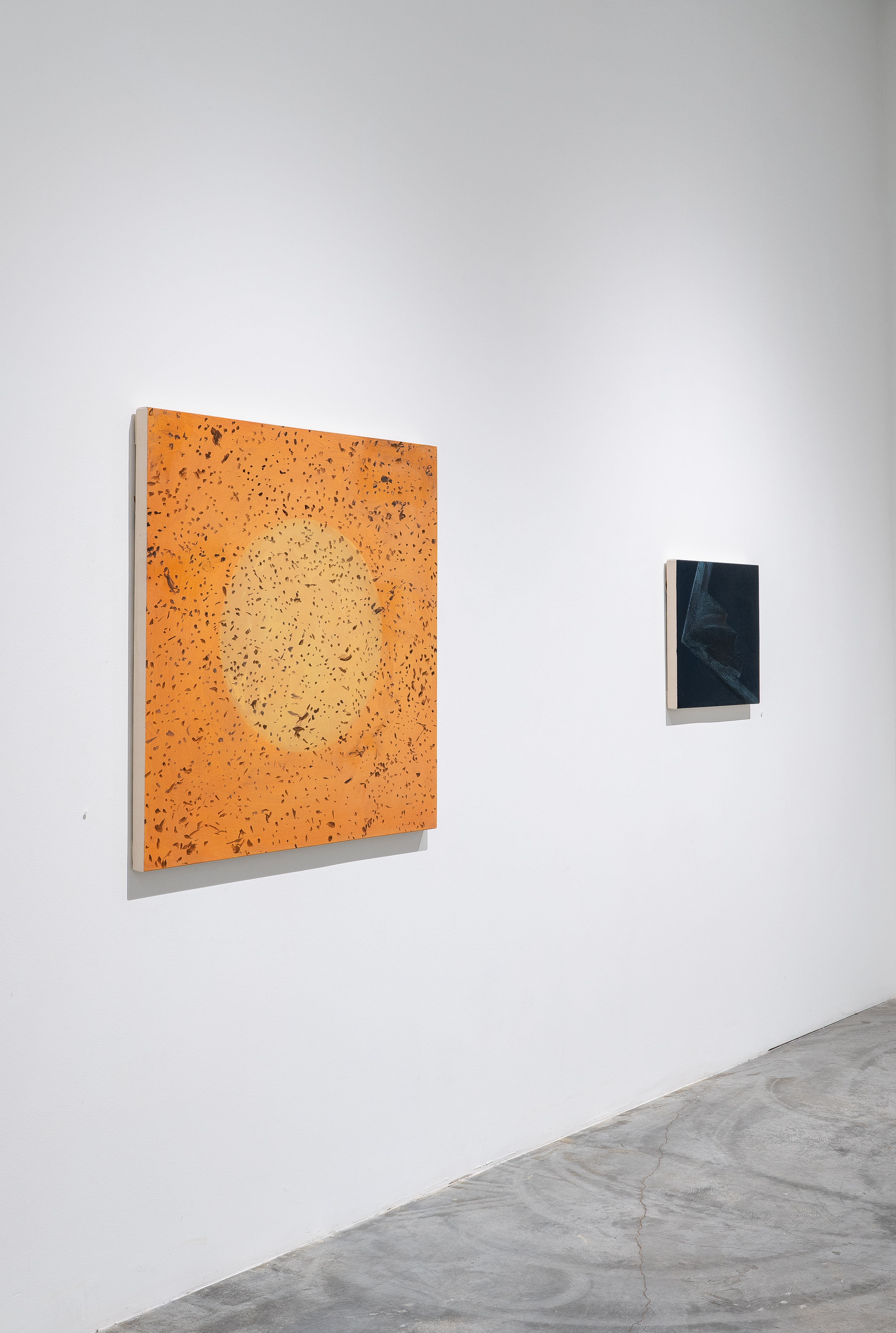 Two abstract paintings mounted on a white gallery wall above a polished concrete floor: a large square composition with warm orange and tan cork or textured material featuring darker speckled patterns, and a smaller rectangular work in dark navy blue with subtle surface variations.