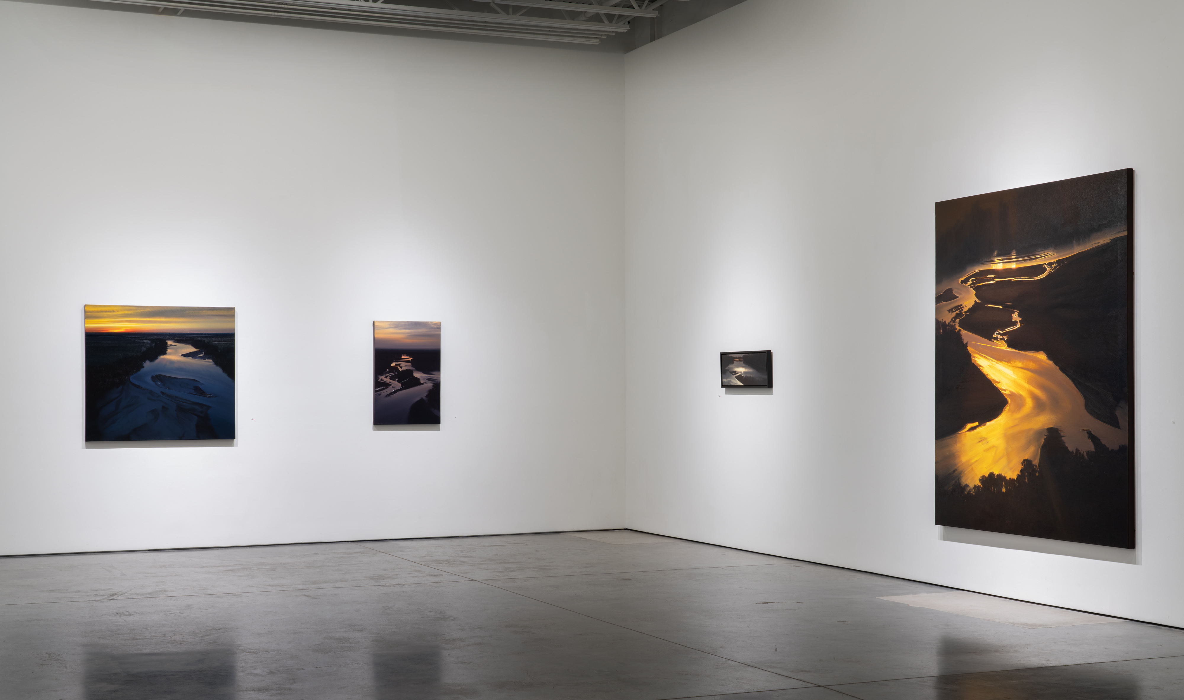 Gallery interior with four framed photographs of aerial river views displayed on white walls. The images depict winding waterways illuminated by golden light against dark landscapes, ranging from small to large scale. Polished concrete floor reflects the gallery space.