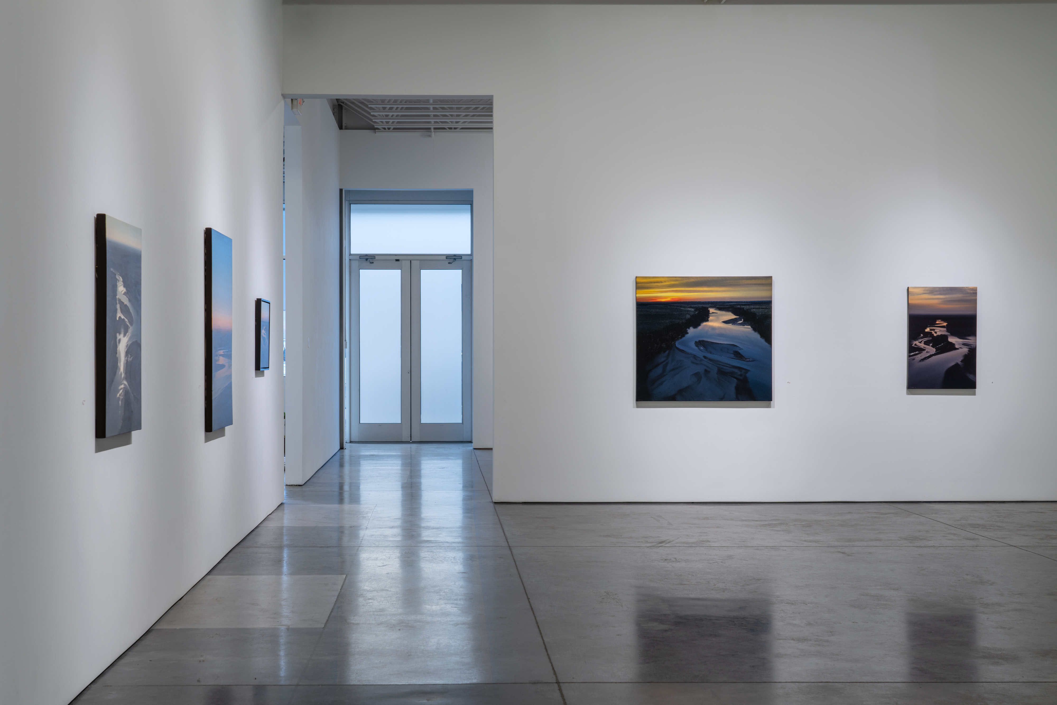 Contemporary art gallery interior with polished concrete flooring and white walls. Several framed photographs depicting aerial views of rivers and landscapes with golden and blue tones hang on the walls at varying heights. A glass door with transom window is visible in the background, allowing natural light into the minimalist space.