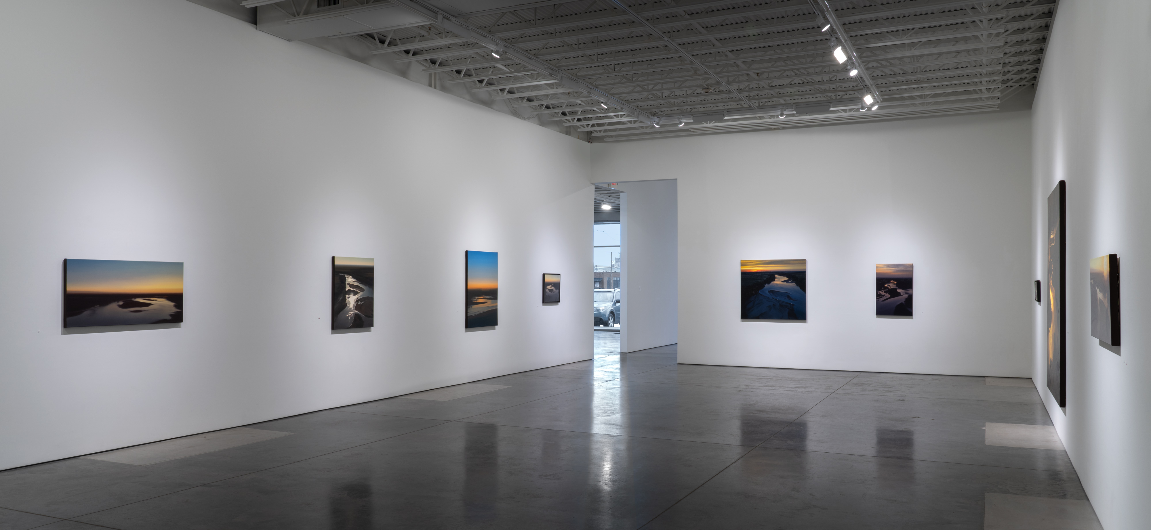 Contemporary gallery space with polished concrete floors displaying aerial landscape photographs mounted on white walls. The images depict winding rivers and waterways photographed from above, rendered in warm sunset tones of orange, gold, and deep blue. Track lighting illuminates the sparse arrangement of framed photographs across the gallery's clean, minimalist interior.