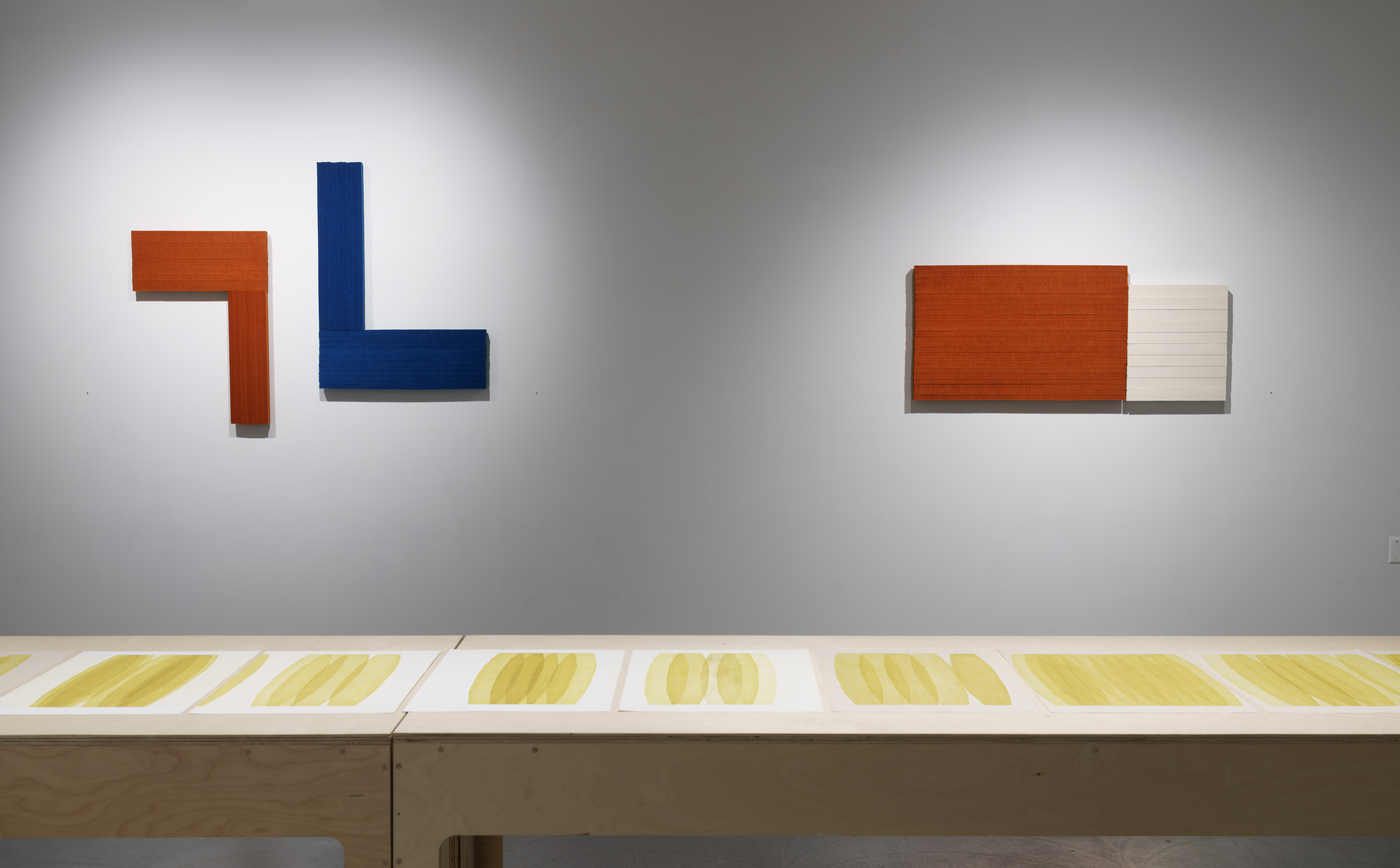 Two wall-mounted sculptural works in contrasting materials: on the left, an angular L-shaped form in rust-orange and deep blue industrial felt with dimensional depth; on the right, a horizontal rectangular composition pairing rust-orange felt with cream-colored panels. Below, a gallery floor displays a series of woodcut prints with golden-yellow oval forms arranged in repeating sequences across white and neutral panels.