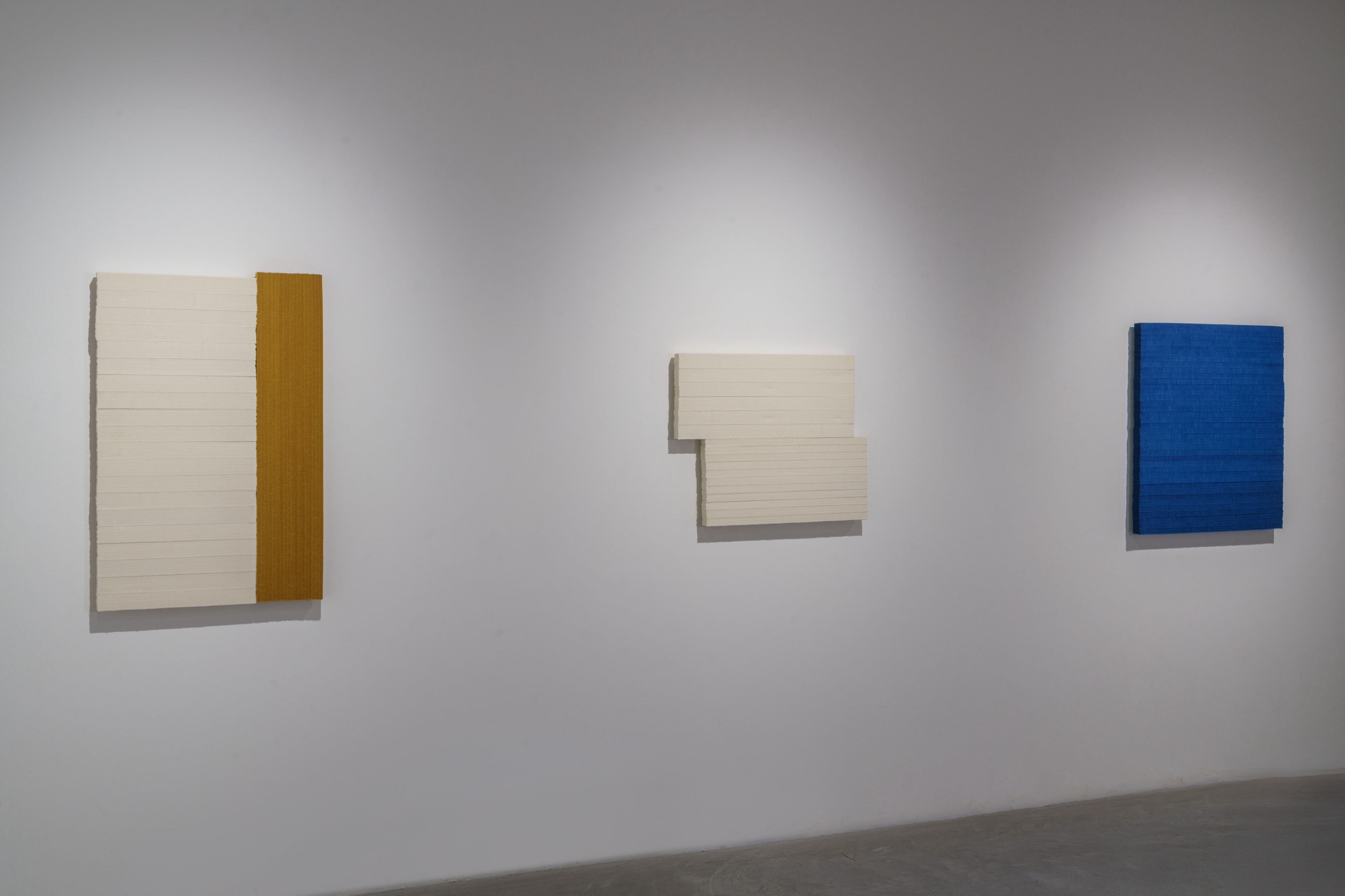 Three wall-mounted abstract compositions in varying scales arranged horizontally against a white gallery wall. Left: cream-colored lined surface with vertical golden-brown wood panel. Center: cream-colored form with geometric cutout creating negative space. Right: deep blue rectangular plane. Each casts a subtle shadow and demonstrates layered, minimalist geometric abstraction.