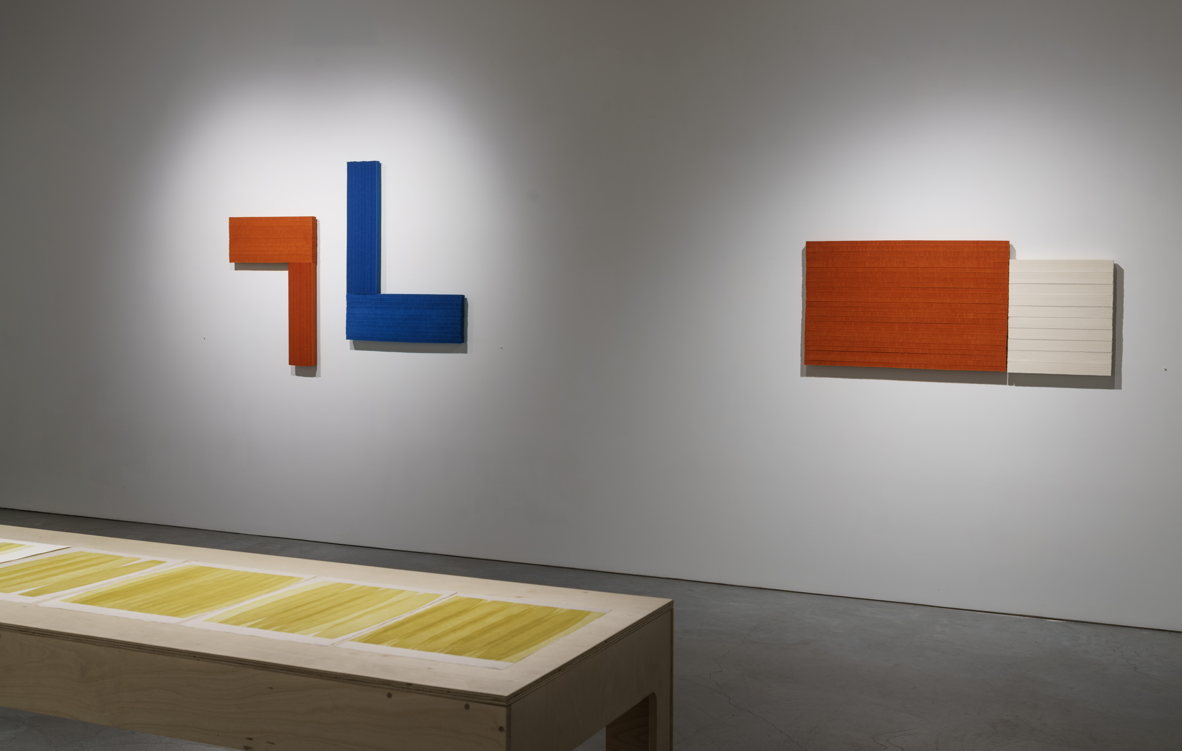 Two geometric abstract wall sculptures in contrasting colors and materials: on the left, an angular L-shaped form in rust orange and deep blue; on the right, a rectangular composition in rust orange and cream. Below, a wooden platform displays a monoprint with yellow and white horizontal striations.