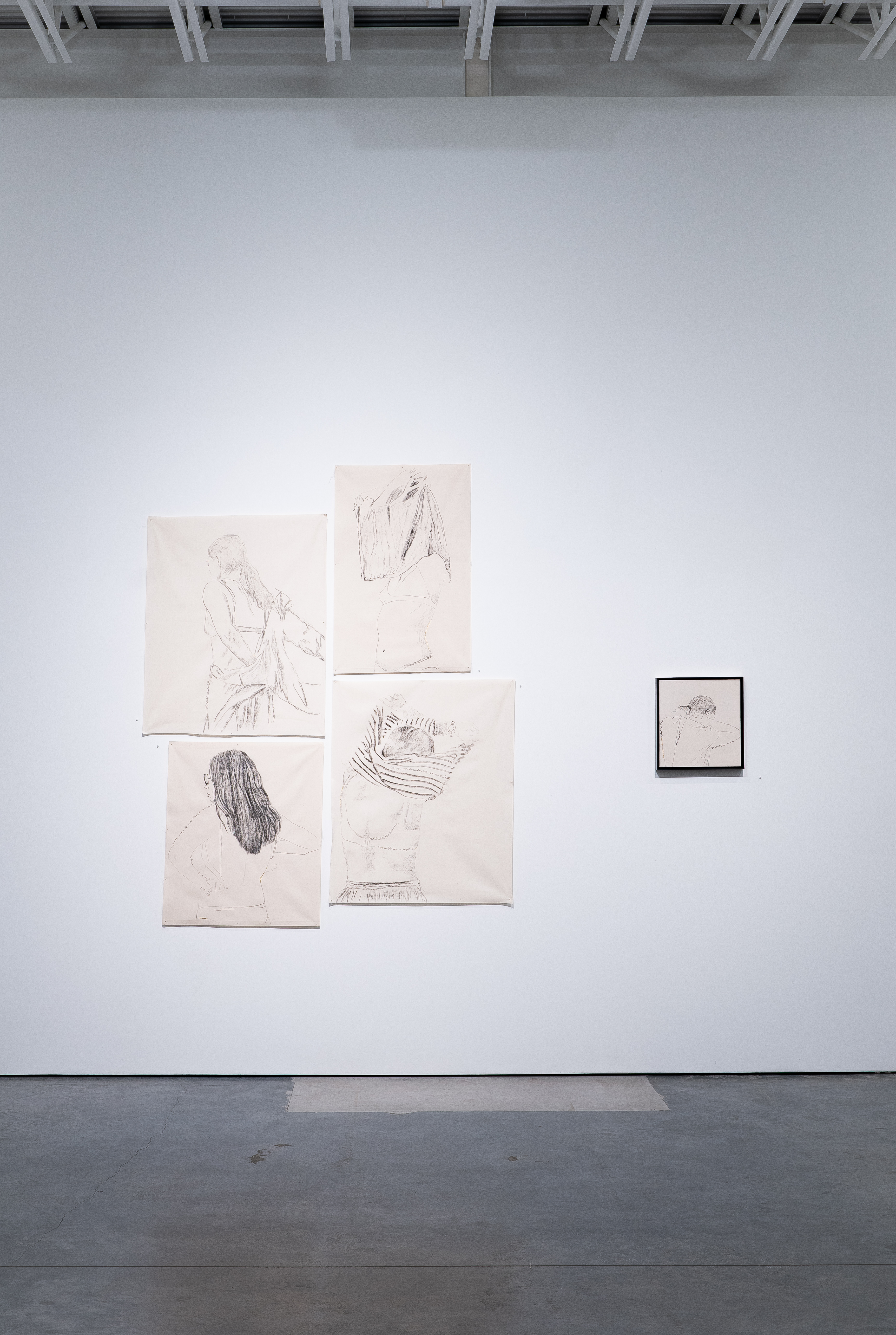 Gallery wall displaying five cream-colored sketches arranged in an asymmetrical cluster, featuring delicate charcoal line drawings of draped fabric and partial figures, alongside a smaller framed drawing mounted separately to the right. The minimalist presentation emphasizes the fragility and intimacy of the gestural marks against the stark white wall.