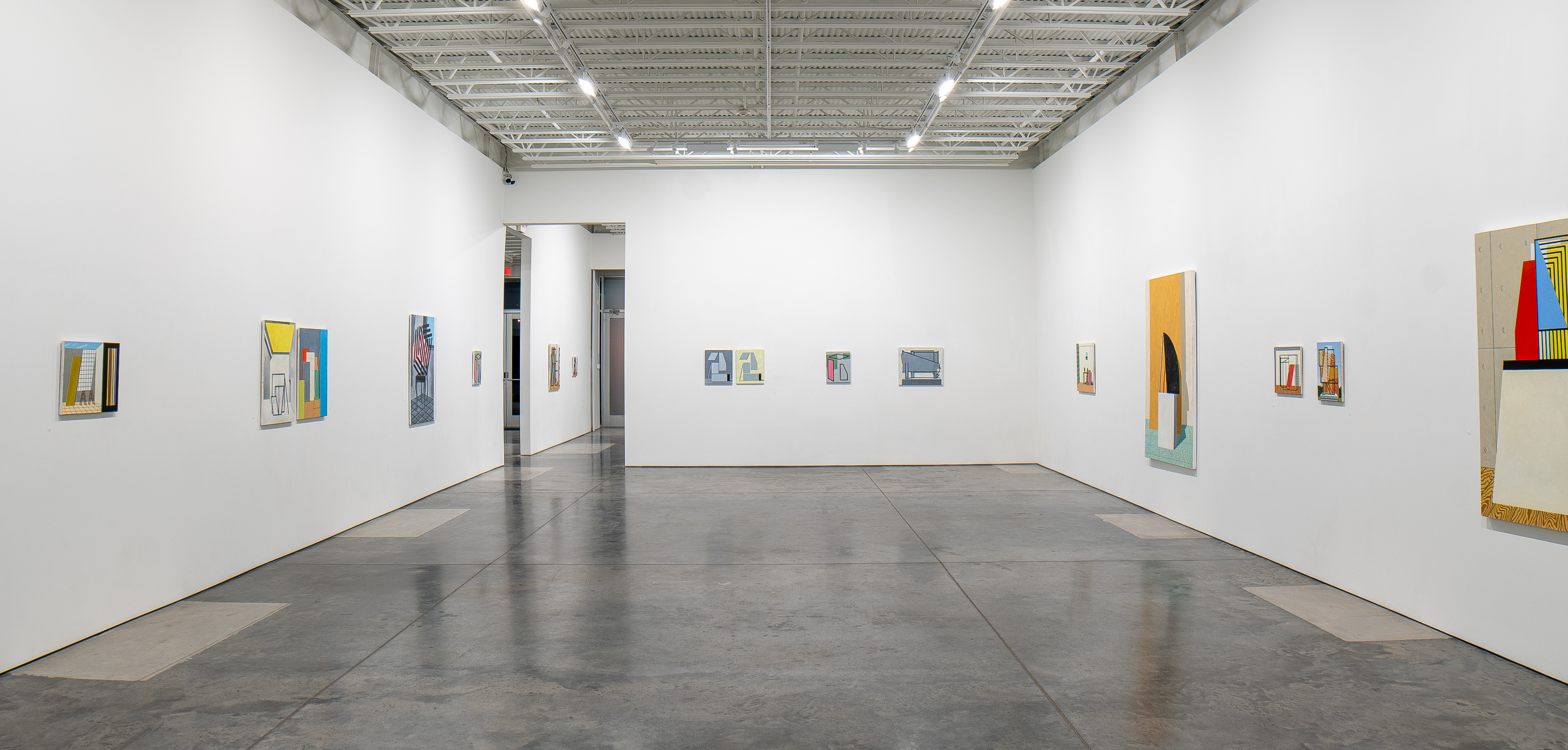 Spacious gallery interior with polished concrete floor displaying approximately fifteen small framed paintings distributed across white walls. The works feature geometric abstraction and architectural references rendered in vibrant colors including yellows, blues, reds, and earth tones, each piece appearing to combine representational and non-objective elements within rectangular frames of varying sizes.