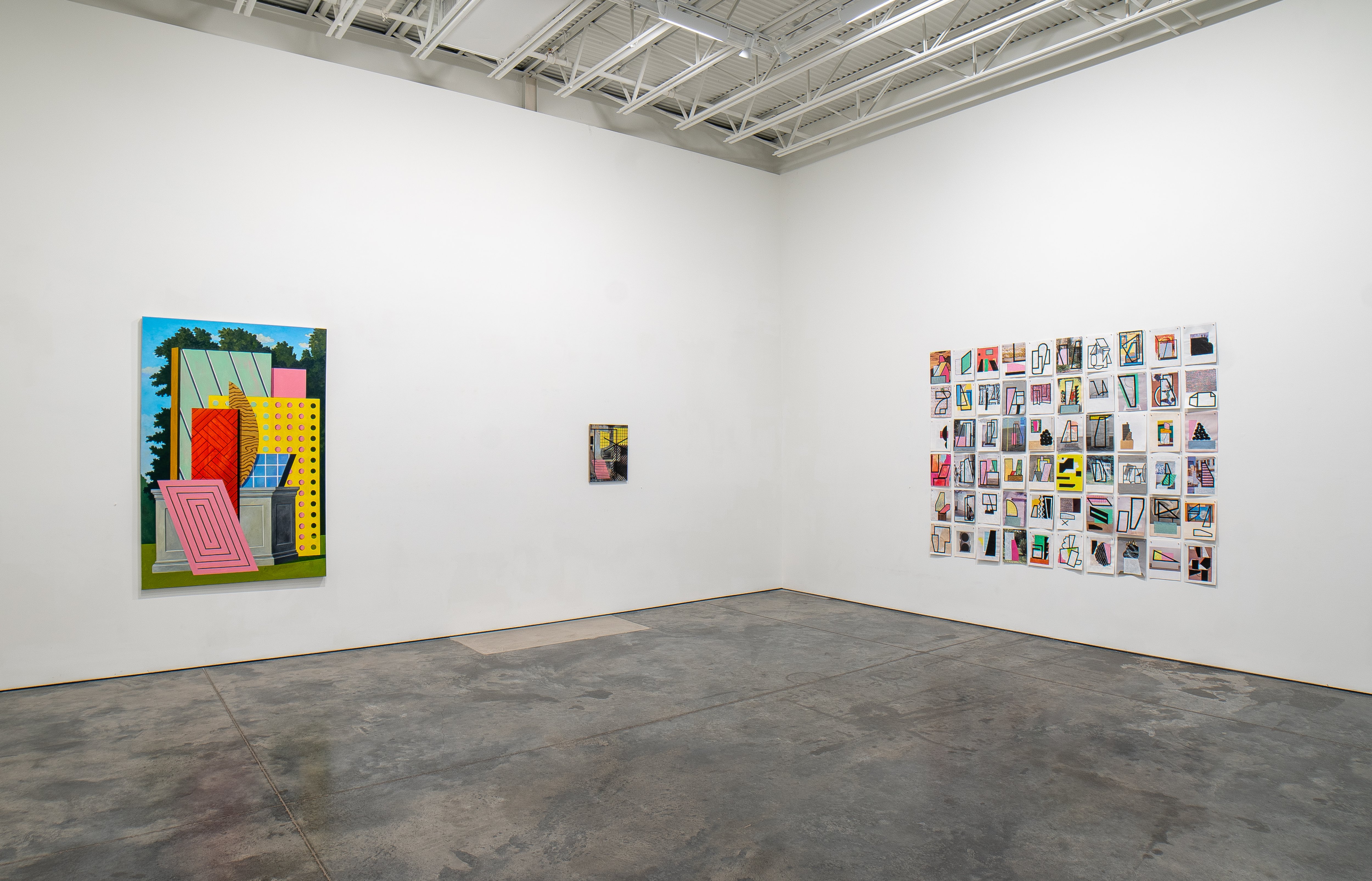 Gallery interior displaying three artworks: a large colorful painting on the left wall featuring geometric shapes in bright hues including pink, yellow, and blue arranged against a landscape background; a small framed work centered on the back wall; and a grid of approximately 60 small rectangular prints on the right wall containing architectural and interior photographs. Polished concrete floor and industrial ceiling with exposed beams.