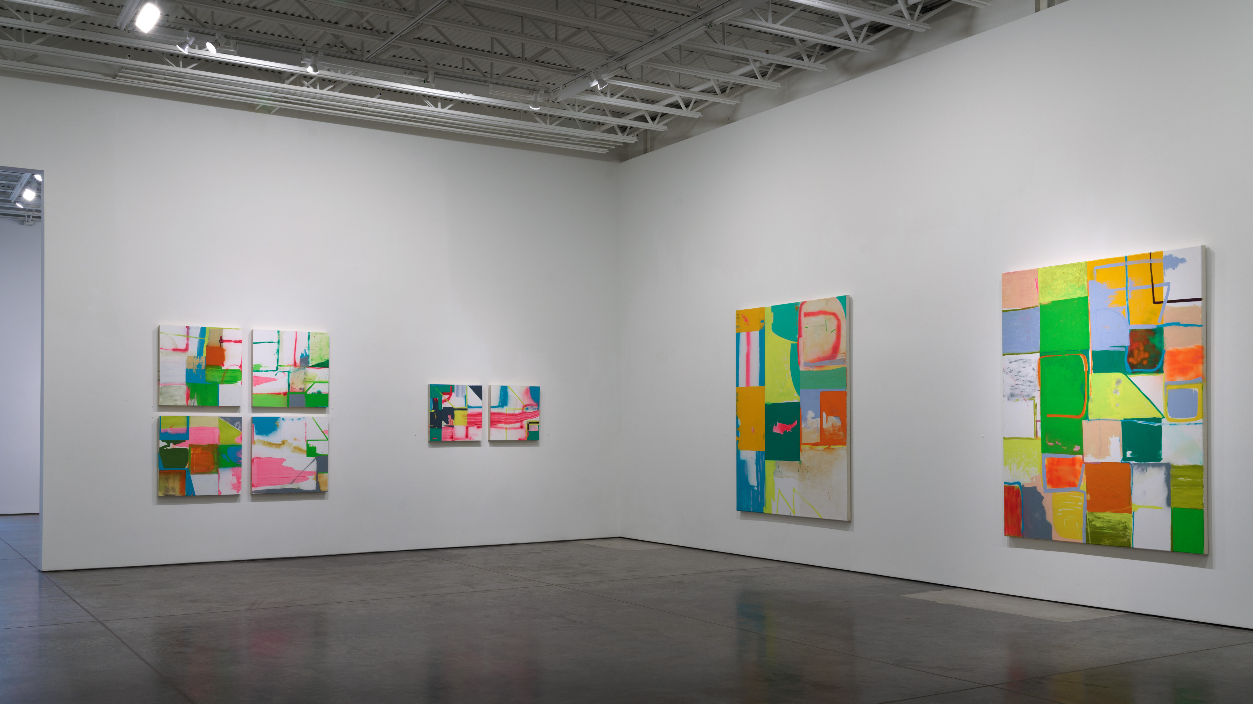 Gallery installation view of four abstract paintings with vibrant color-block compositions arranged on white walls. Each work features geometric grids of rectangles in bright hues—greens, yellows, oranges, pinks, and blues—interspersed with white negative space, creating an open, airy aesthetic across the polished concrete floor.