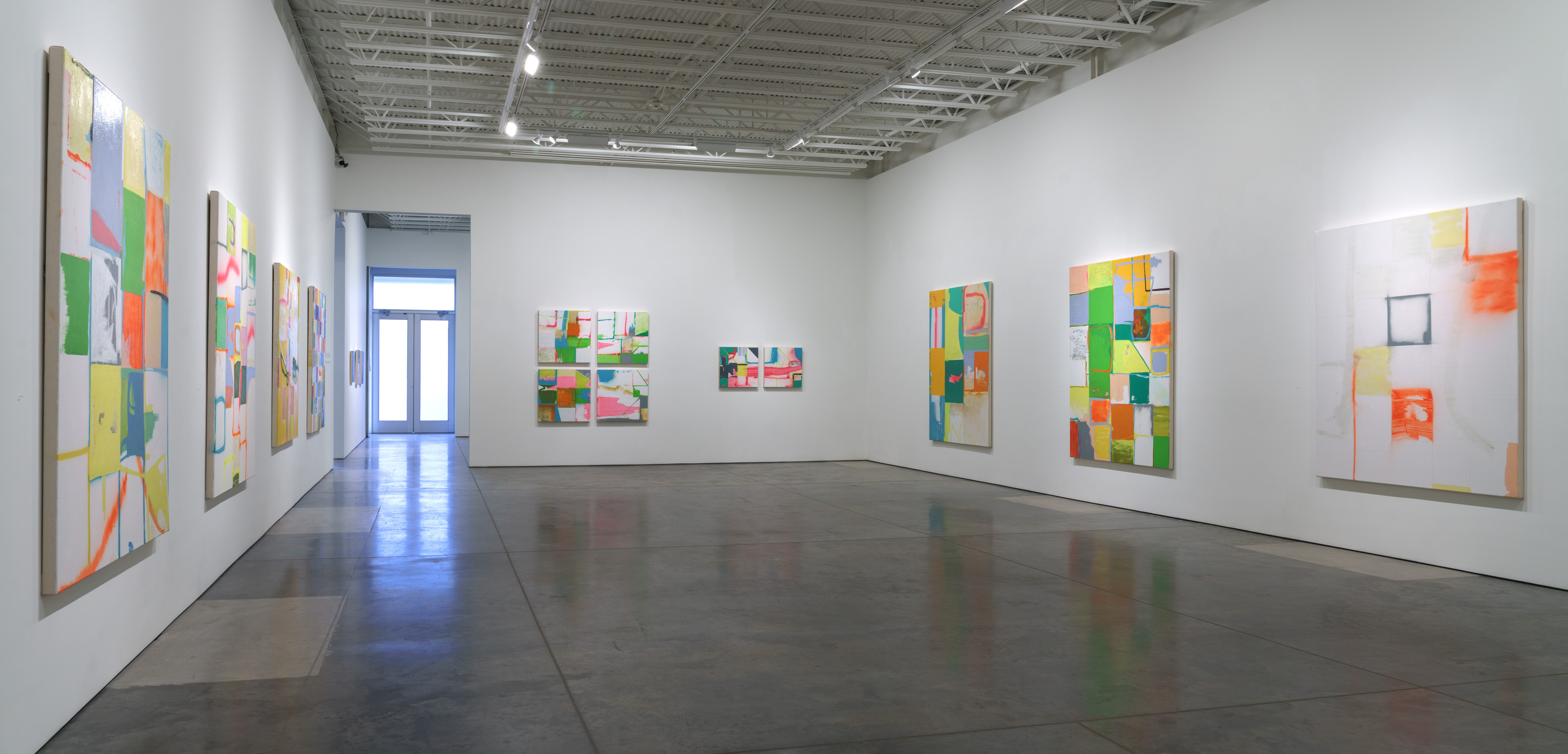 Gallery interior displaying a series of colorful abstract paintings on white walls. The works feature vibrant grids and geometric compositions in shades of green, yellow, orange, blue, and pink, with generous areas of white space. Paintings of varying sizes are arranged across the gallery's pristine walls, with polished concrete flooring reflecting the bright overhead lighting from an industrial ceiling.