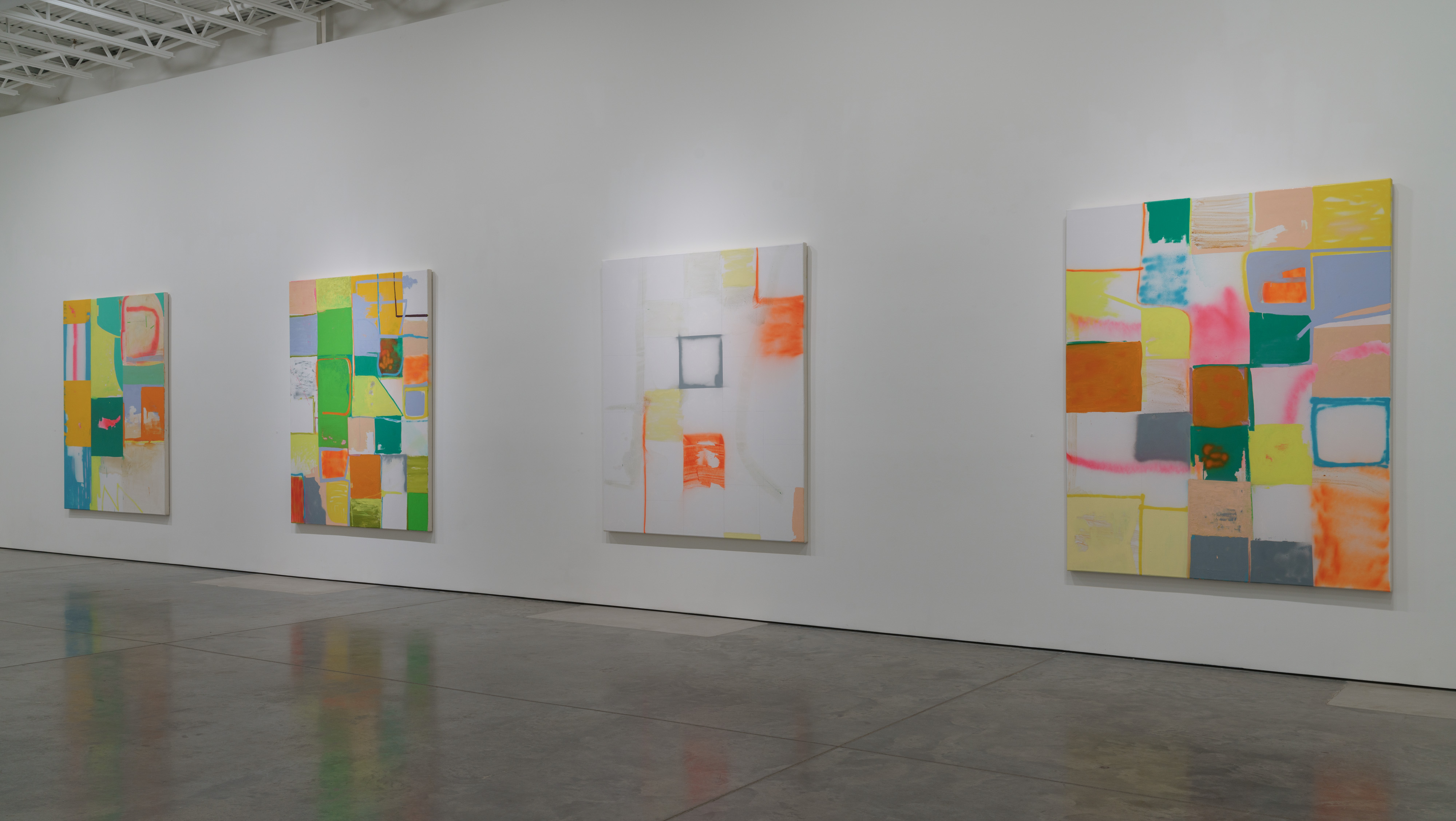 Four colorful abstract paintings displayed on white gallery walls featuring geometric grids composed of rectangular shapes in vibrant yellows, oranges, greens, blues, and pinks interspersed with areas of white space and outlined rectangles, arranged in a sparse, open composition against a polished concrete floor.