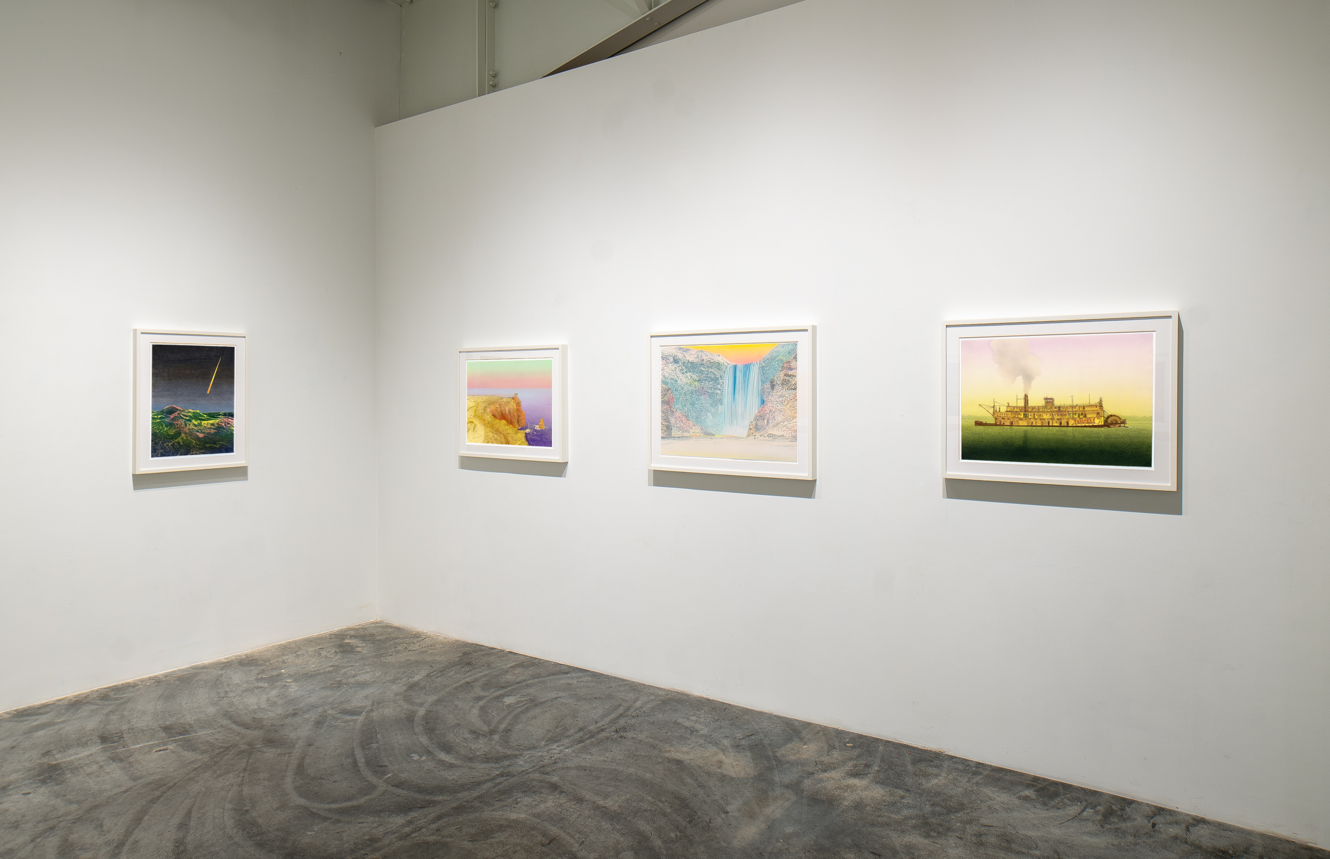 Four framed landscape photographs arranged horizontally on a white gallery wall above a polished concrete floor. From left to right: a dark nocturnal scene with a glowing yellow streak, a colorful cliff or rock formation at sunset, a misty waterfall cascading down layered rock faces, and a golden-hour view of an ornate building silhouetted against a luminous sky. Each print is mounted in a white frame with a gray shelf-like base.