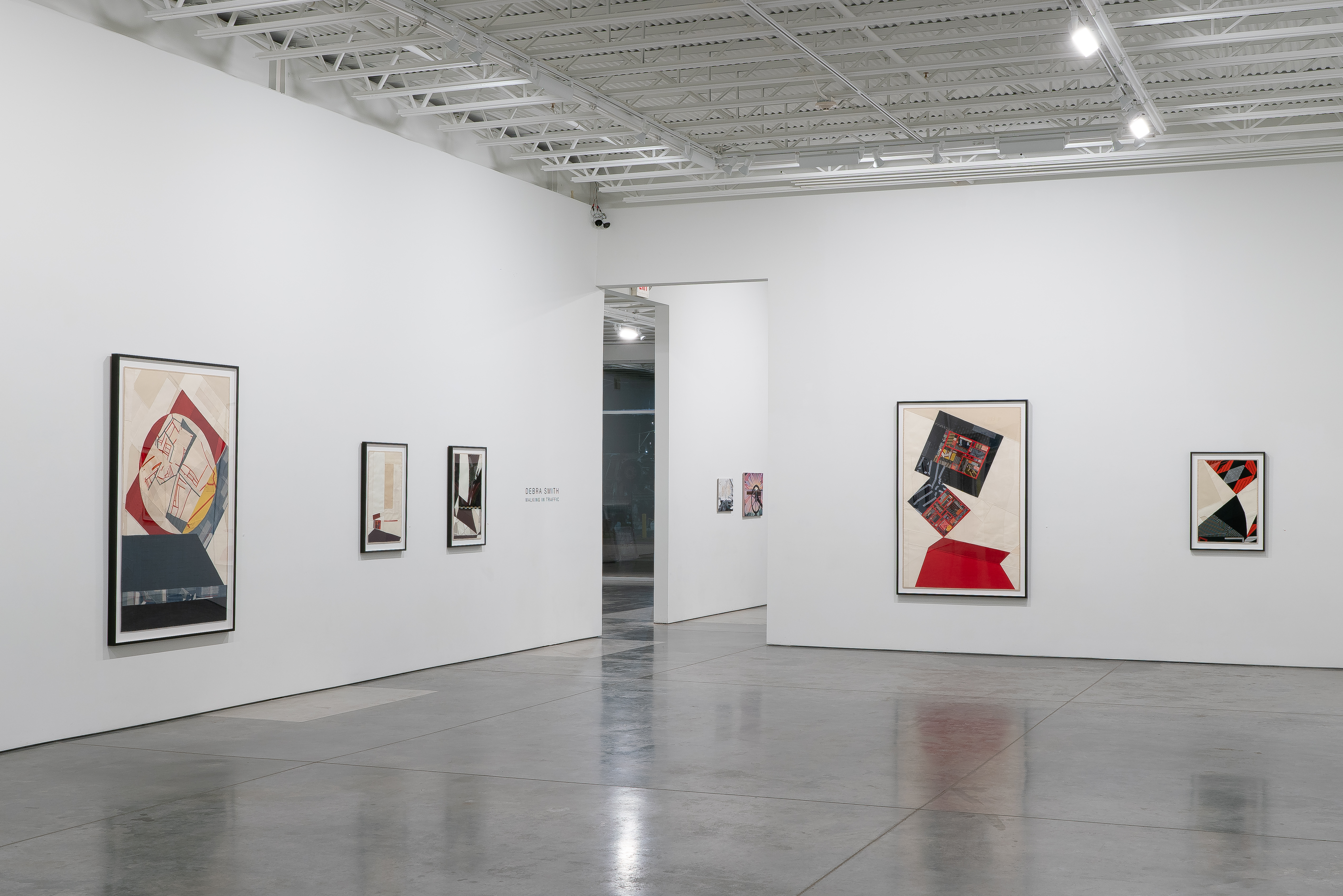 Contemporary gallery interior displaying framed textile collages on white walls. The works feature bold geometric compositions with vibrant red, black, and cream-colored vintage fabrics arranged in abstract patterns. Pieces vary in scale from small to large formats, arranged across the bright, minimalist space with polished concrete flooring and industrial ceiling.
