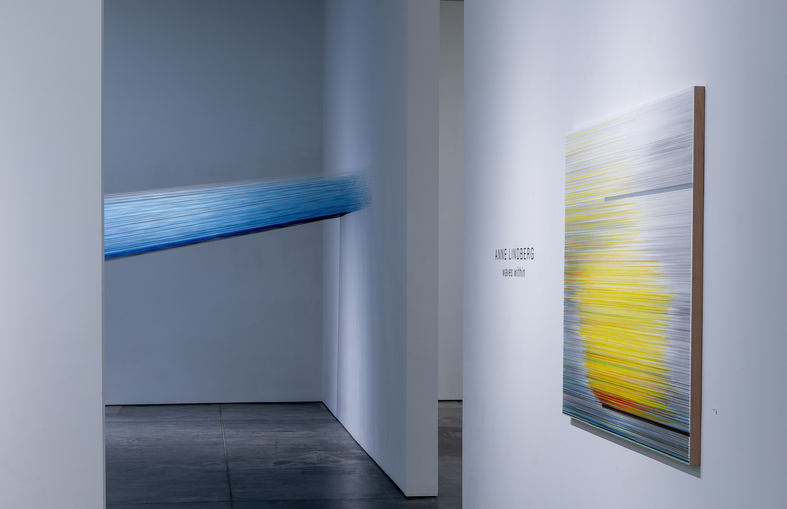 A gallery interior with white walls and dark flooring features two artworks: a large-scale installation of taut indigo-blue threads creating a luminous gradient that stretches horizontally across the left wall, and a vertical work on the right wall composed of horizontal bands of yellow, blue, gray, and orange lines creating a wave-like pattern. The gallery space is minimalist and brightly lit.