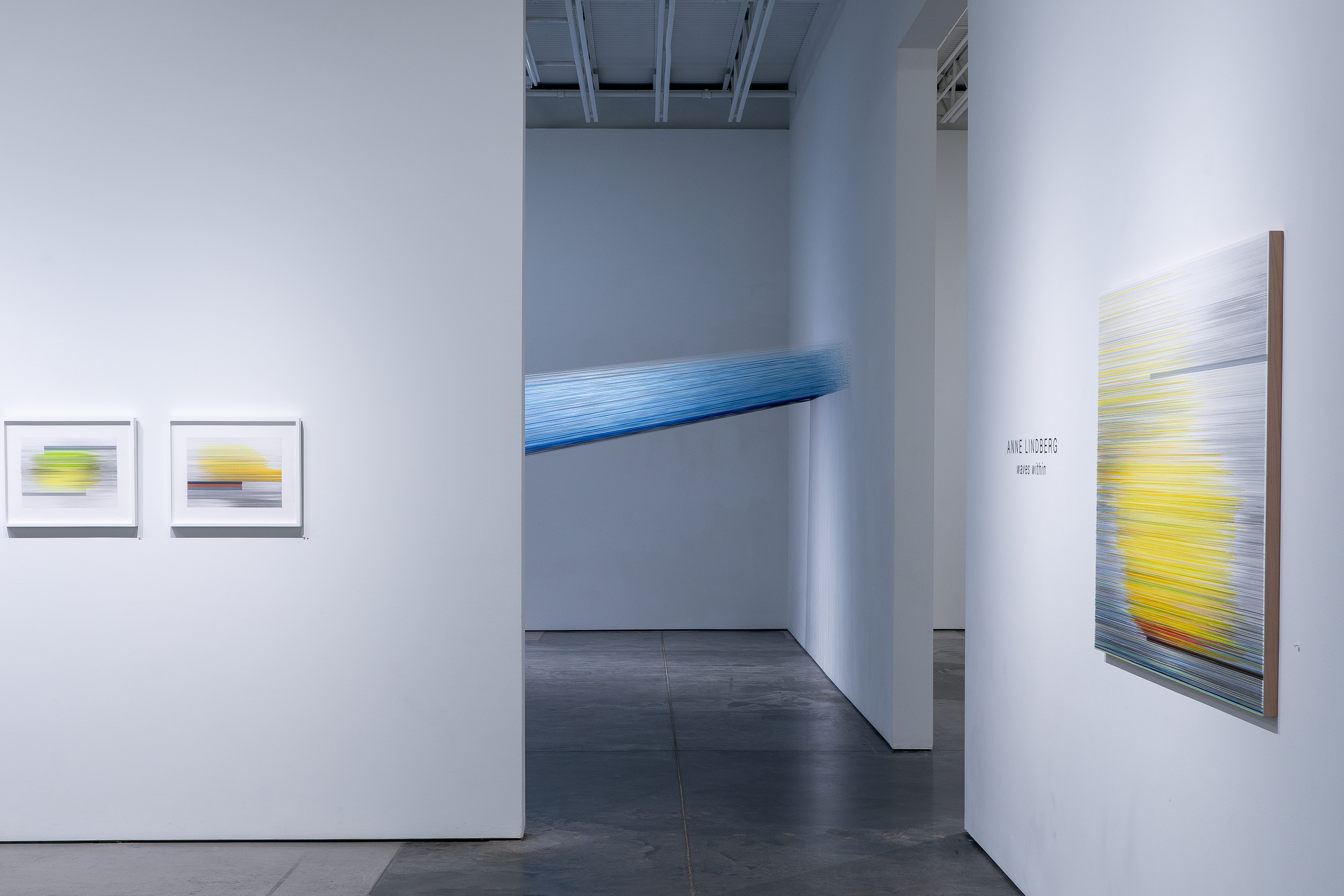 Contemporary gallery installation featuring framed works with horizontal striations in yellow, green, and blue tones on the left walls, and a large-scale immersive installation of taut indigo-dyed threads suspended diagonally across the central gallery space, creating a luminous gradient effect throughout the room.