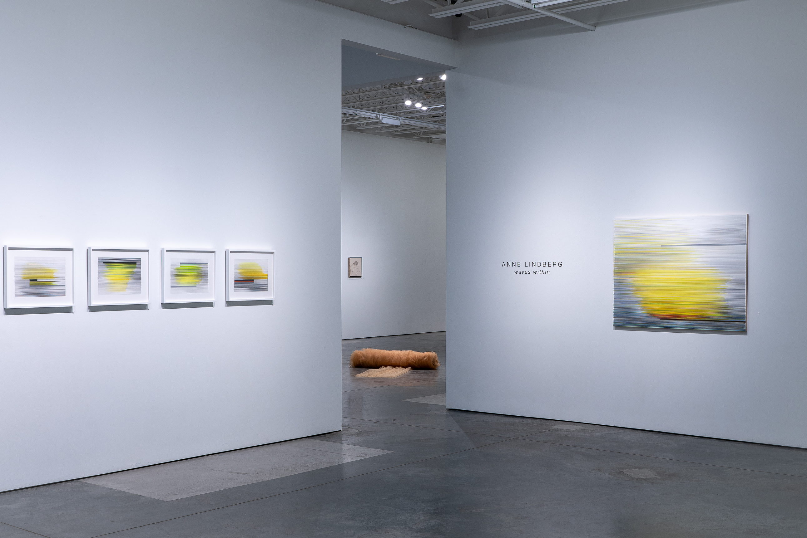 Contemporary gallery installation featuring four framed drawings with yellow and gray horizontal striations on the left wall, a larger unframed drawing with similar imagery on the right wall, and a sculptural element visible through a doorway. The works employ gestural marks and color shifts suggestive of landscape and atmospheric conditions.