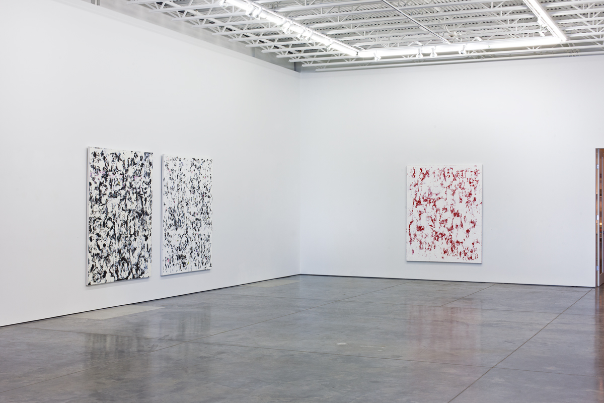 A gallery space displaying three large abstract paintings on white walls. Two black and white gestural works hang on the left wall, while a red and white abstraction occupies the right wall. Each canvas features dense, layered brushstrokes and organic marks across the surface. The polished concrete floor reflects the gallery's industrial ceiling with exposed metal trusses.