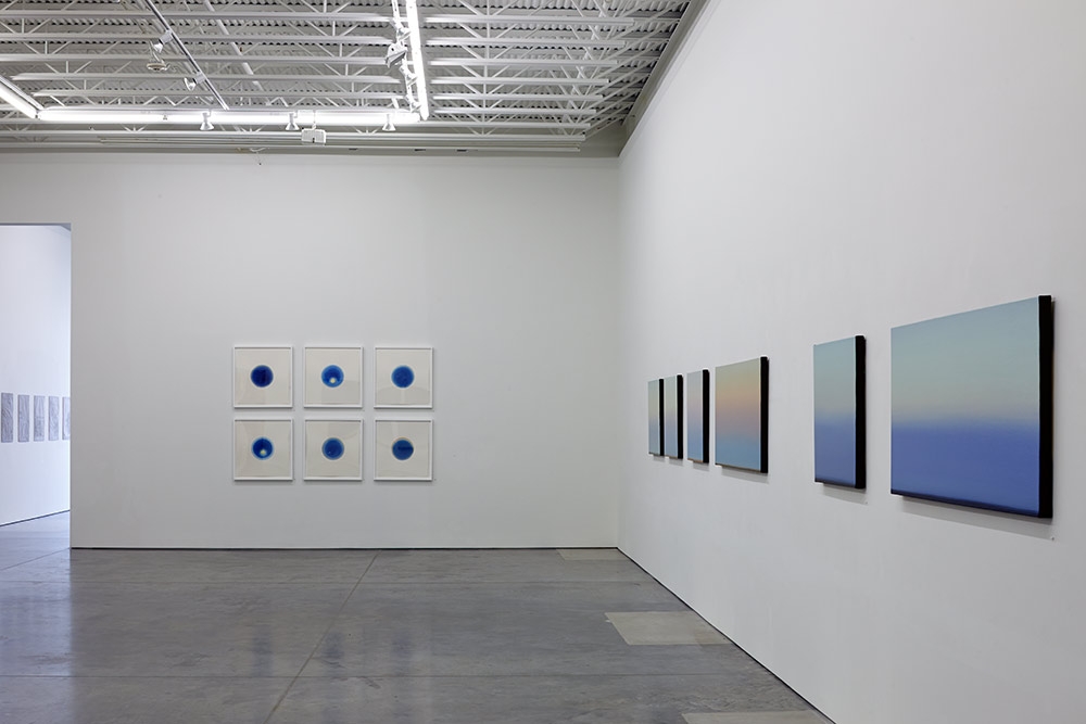 Contemporary gallery interior with white walls and industrial ceiling. On the left wall, six small framed works arranged in a 2x3 grid, each featuring a deep blue circular form on a light background. On the right wall, a series of larger horizontal paintings with gradient compositions transitioning from warm peachy tones at the top to deep blue at the bottom, mounted with black frames.