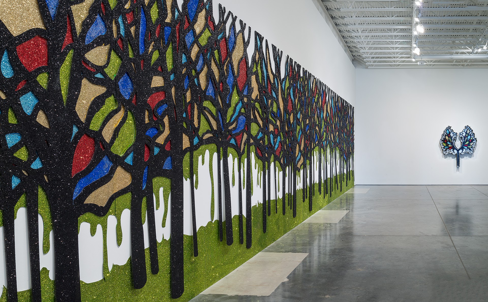 A large-scale wall mural featuring an inverted forest of black trees with colorful geometric shapes in red, blue, lime green, and gold glitter covering the canopy against a dark speckled background. White dripping forms cascade down the tree trunks onto a bright green ground, with a single jeweled ornamental tree sculpture mounted on the adjacent white gallery wall.