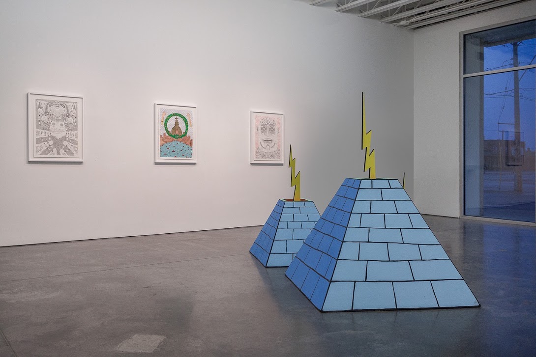Two blue pyramidal sculptures with yellow lightning bolt spires stand on a polished concrete floor in a gallery space, accompanied by three framed drawings on the white wall behind them featuring intricate line work and symbolic imagery.