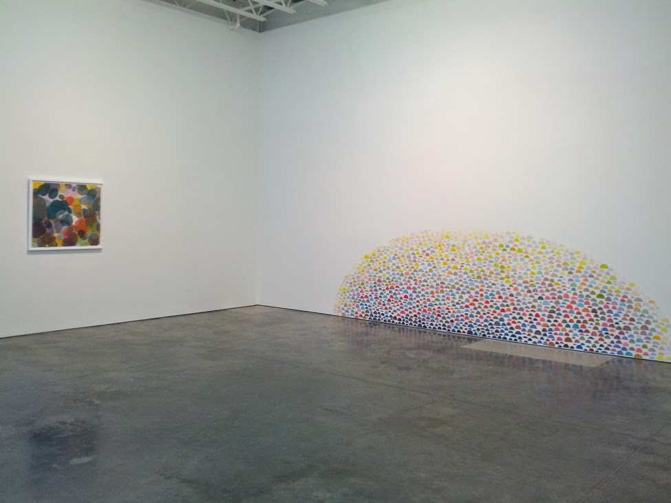 A gallery space displaying a small framed abstract print with colorful circular patterns on the left wall, and a large dome-shaped sculptural installation on the floor composed of thousands of tightly packed colored spheres in yellow, white, blue, red, and green tones, creating a dense, bulbous form that appears to emerge from or rest against the corner.
