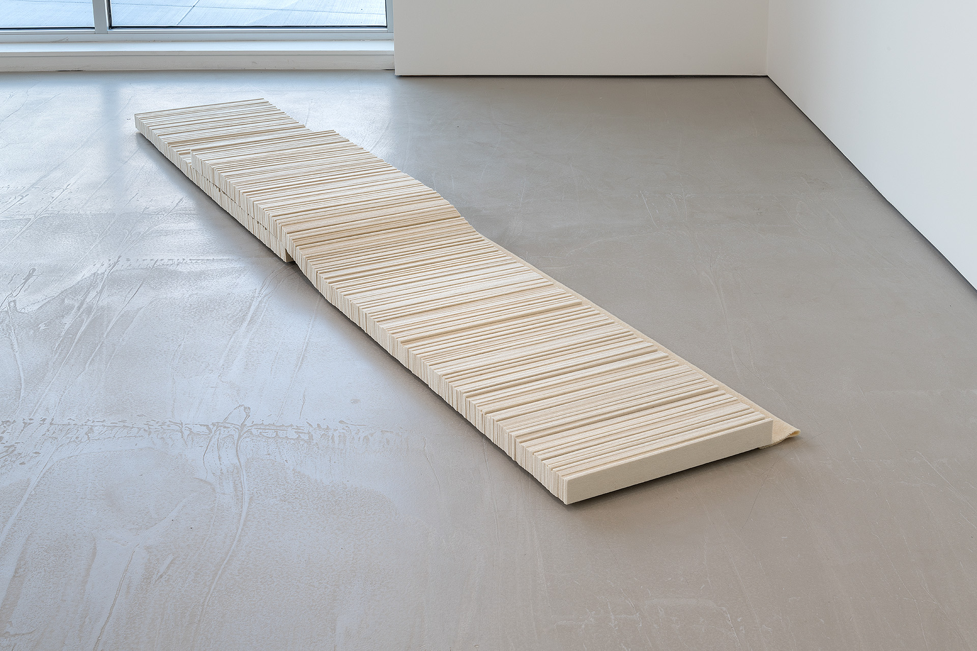Long wooden sculptural form with horizontal linear striations lying diagonally across a concrete gallery floor, positioned between white walls and a window, evoking architectural minimalism through stacked and folded natural materials.