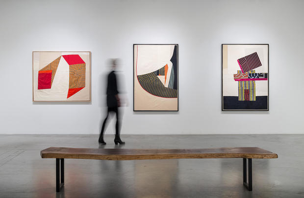Three framed textile compositions hang on a white gallery wall, each featuring geometric abstract designs created from hand-cut pieces of patterned and solid-colored fabric in red, orange, black, and neutral tones. A wooden bench sits in the foreground with a blurred figure standing beside it, viewing the work.