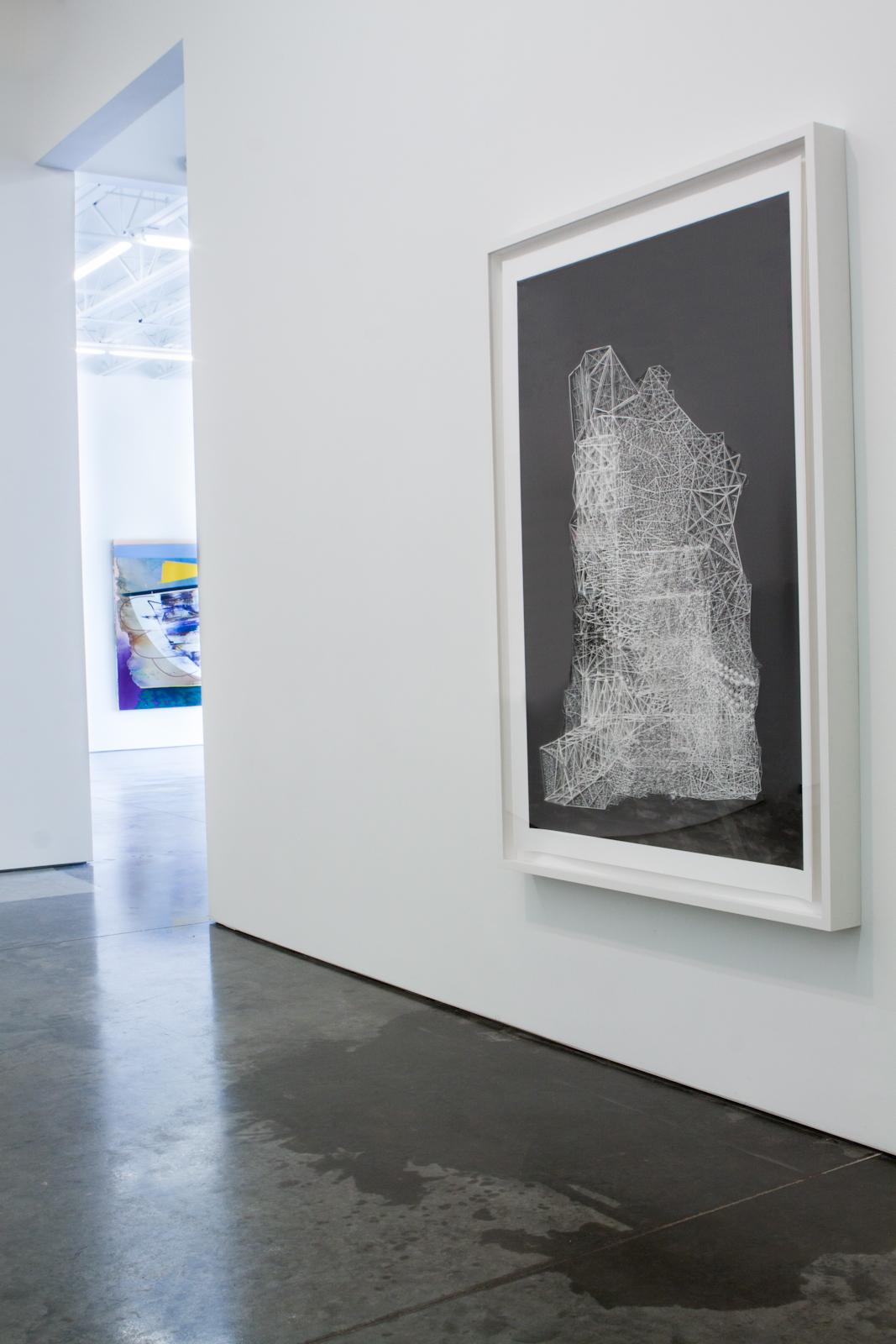 White line drawing of an abstract architectural or geological form against a black background, displayed in a white frame on a gallery wall. The minimalist linear composition contrasts sharply with the dark interior space and polished concrete floor.