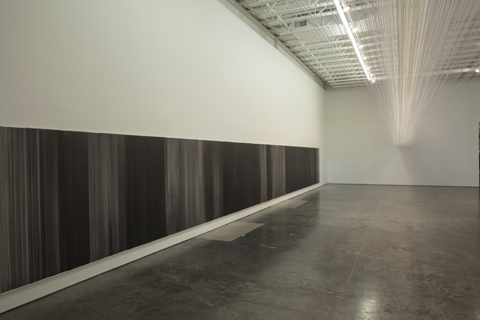 A minimalist gallery installation featuring a dense horizontal band of black vertical lines at eye level along the lower wall, with an ethereal canopy of fine white threads suspended above, creating a contemplative spatial experience in a concrete-floored industrial gallery.