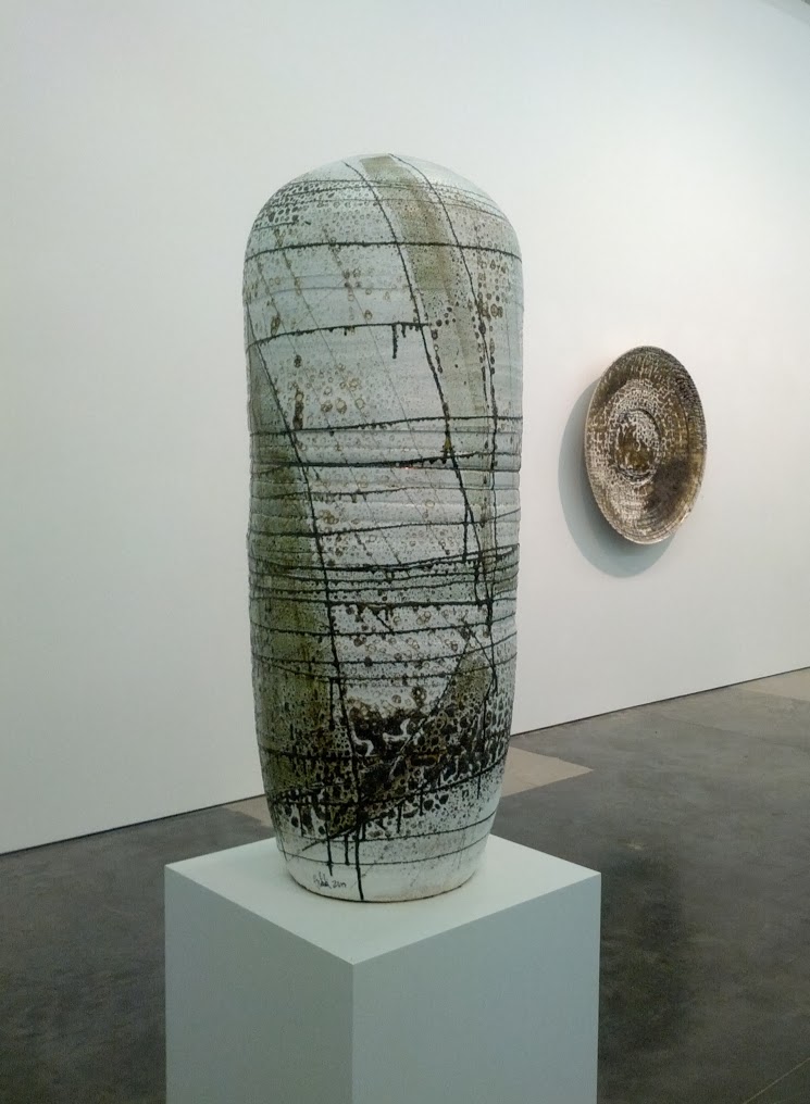 A tall cylindrical ceramic vessel with a cream and muted green surface, covered with horizontal linear markings and darker gestural marks concentrated at the base. The sculpture is displayed on a white pedestal alongside a smaller rounded ceramic disc mounted on the wall, both works suggesting a dialogue between form and negative space.
