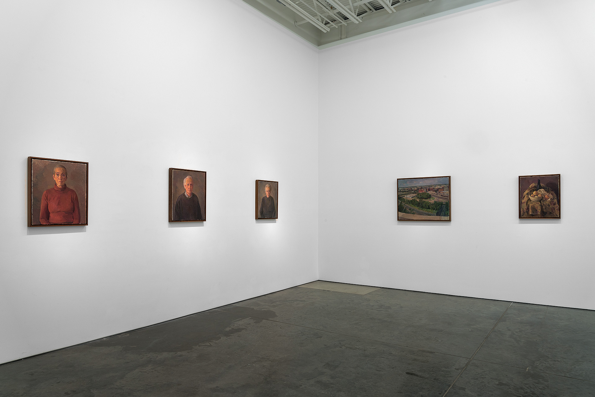 Gallery wall displaying five framed paintings arranged horizontally: three portrait studies of male figures in dark tones on the left, a landscape depicting a hillside town in the center-right, and a still life composition with nude figures on the right. Clean white walls and polished concrete floor create a minimalist exhibition space.
