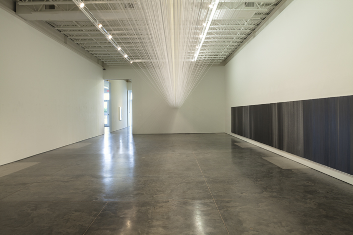 A minimalist gallery installation features a suspended cloud of delicate white threads spanning the ceiling's length, while a dense horizontal band of dark charcoal or black drawing runs along the lower wall, creating a symmetrical composition that activates the expansive concrete floor and emphasizes the viewer's physical presence in the space.