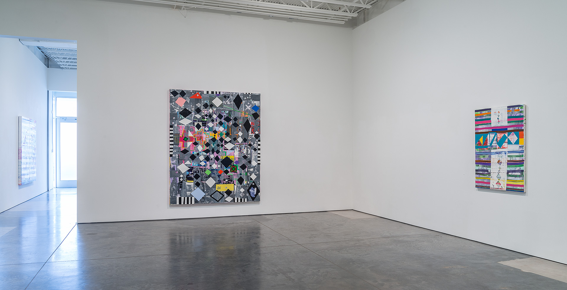 A large abstract painting with a geometric diamond pattern in black, white, and gray dominates the center wall, layered with colorful collaged elements and striped motifs. To the right, a smaller mixed-media work features horizontal bands of vibrant rainbow colors interspersed with geometric shapes and white sections.