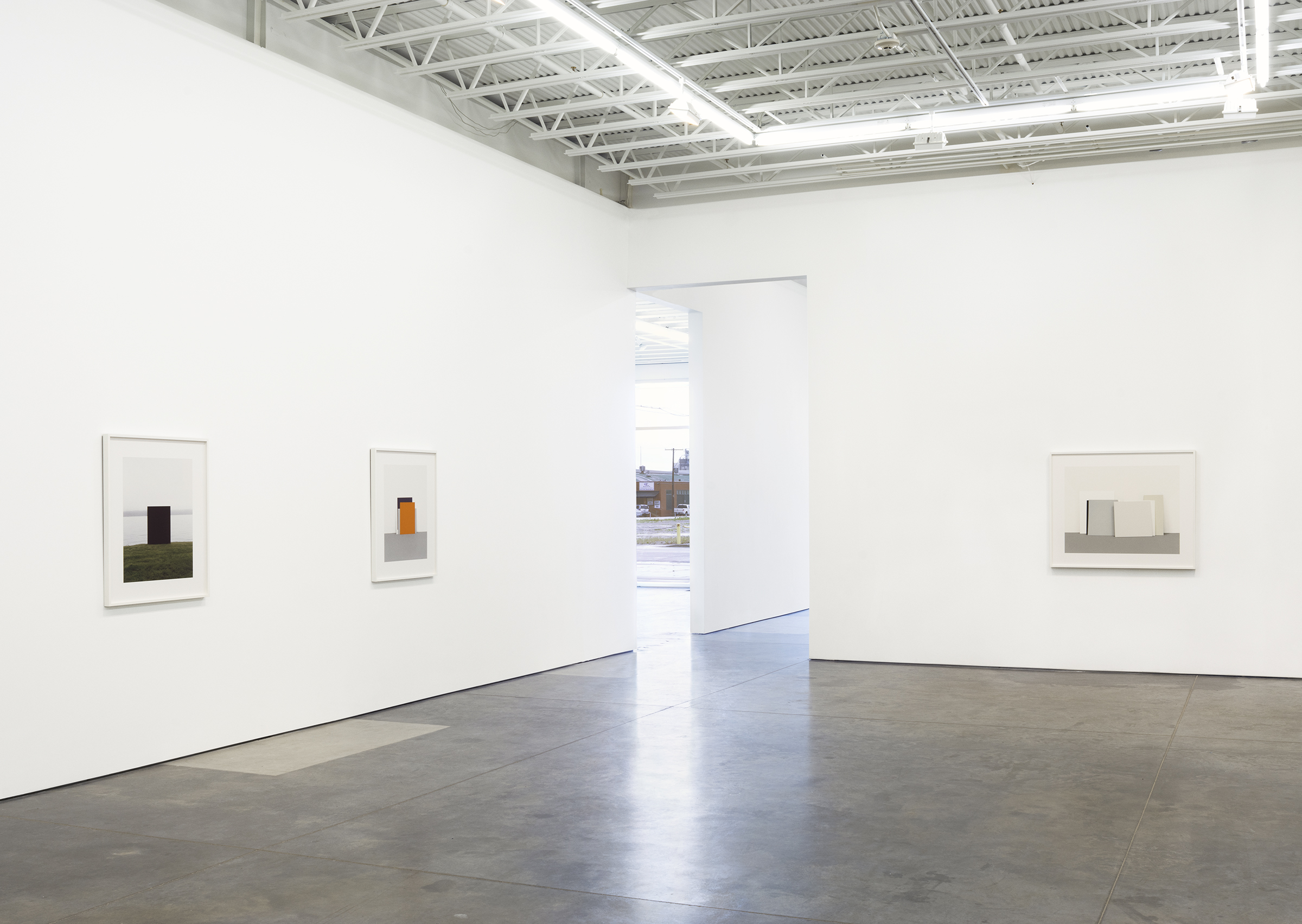 Contemporary gallery space with polished concrete floor and white walls featuring four framed photographs spaced across the walls. Each photograph depicts geometric forms—colored rectangles and blocks—positioned within architectural or landscape settings. Natural light streams through a central opening, illuminating the minimal, clean composition of the gallery interior.