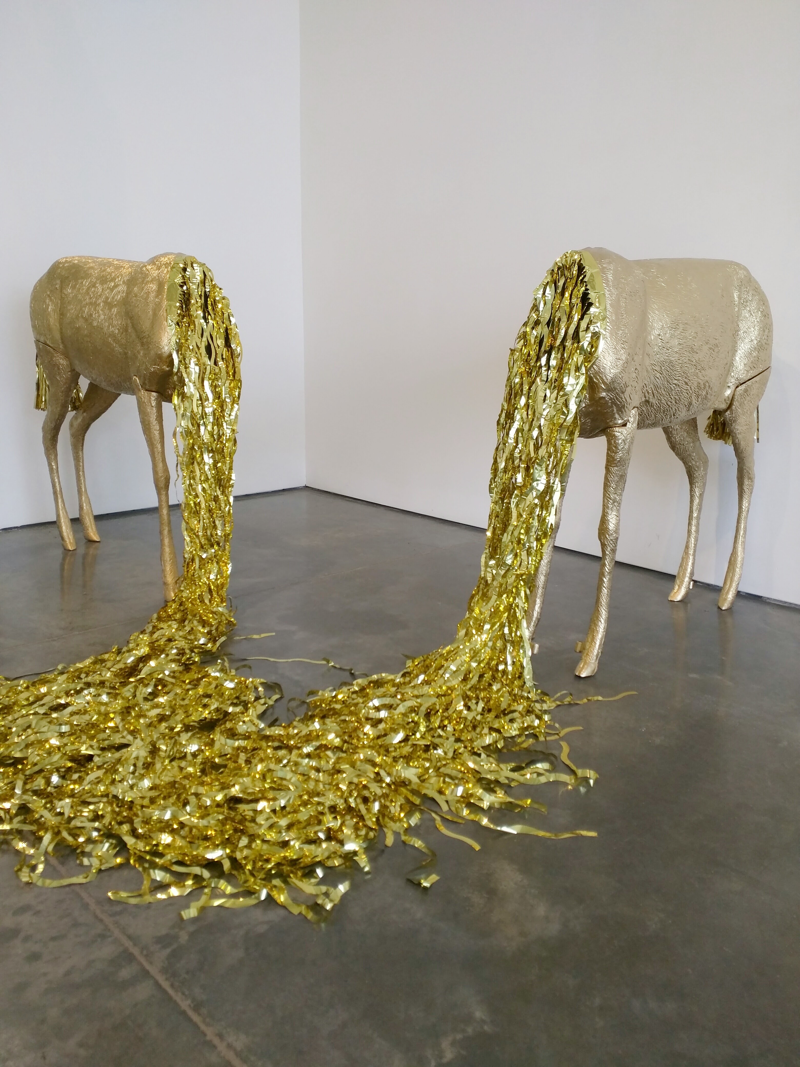 Two pale resin deer figures stand facing away from each other on a dark floor, their heads bowed downward. Cascading from each deer's mouth is an elaborate stream of crinkled gold lamé fabric pooling across the gallery floor, creating a striking contrast between the naturalistic animal forms and the luxurious, reflective material.