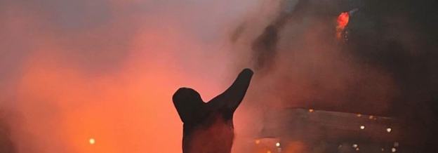 Silhouetted figure with raised arm against a dramatic backdrop of orange and red flames and smoke, rendered in oil painting with gestural brushwork that emphasizes the intensity of the scene.