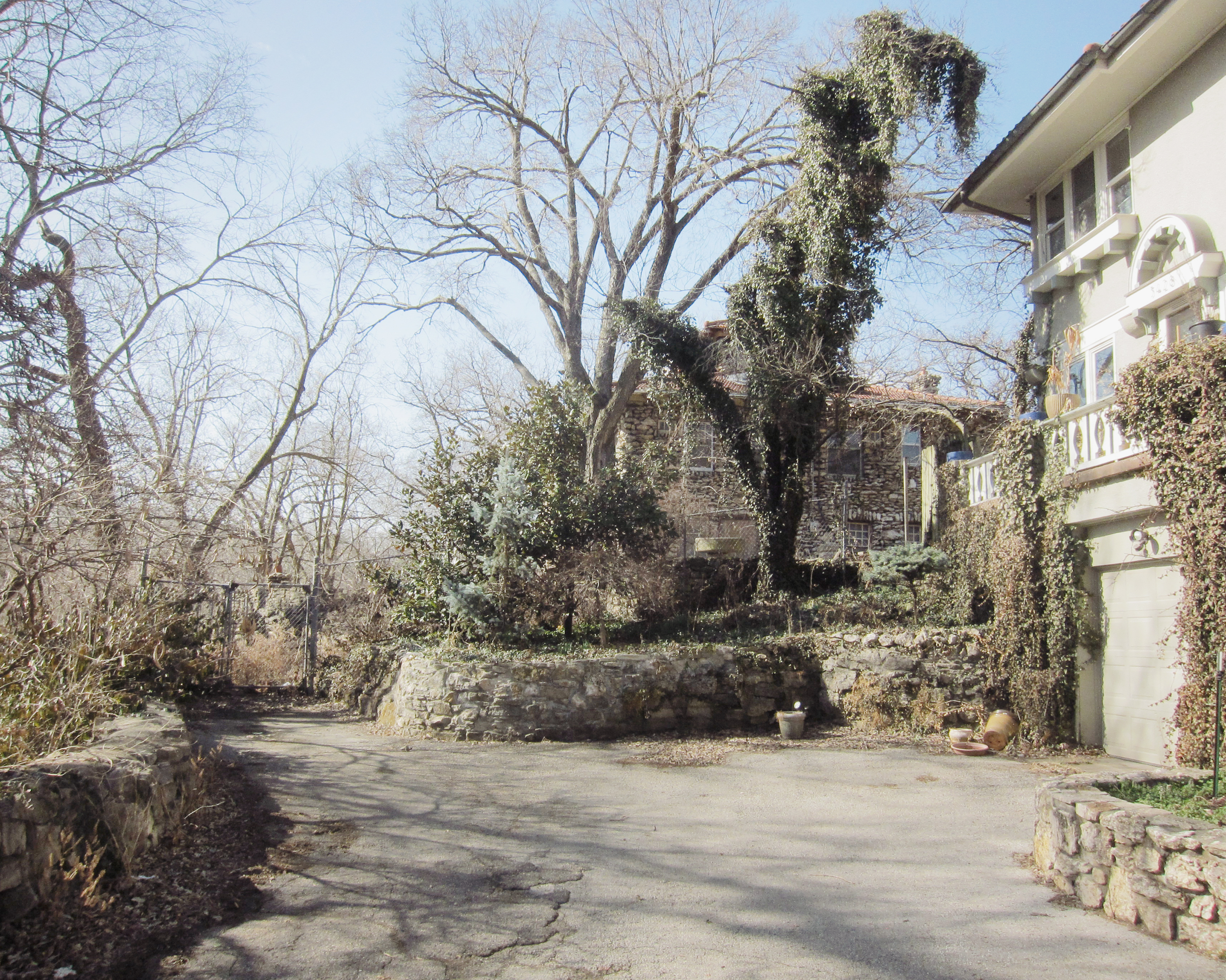 A sunlit courtyard bordered by ivy-covered stone and white buildings, with bare winter trees rising behind weathered walls and a paved ground casting sharp shadows.
