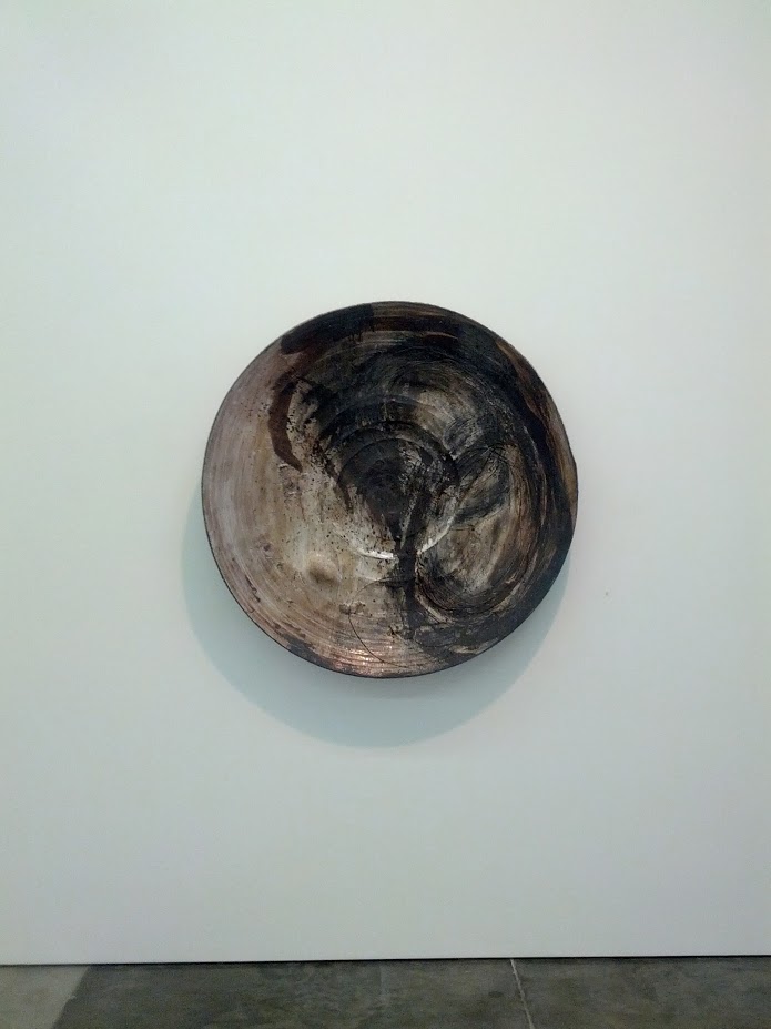 Monumental circular ceramic vessel mounted on a white wall, its concave interior surface revealing layered brushstrokes in charcoal black and gray tones that create a spiraling, meditative composition. The exterior rim transitions from lighter taupe to darker charcoal, with visible gestural marks suggesting both contemplative abstraction and organic form.