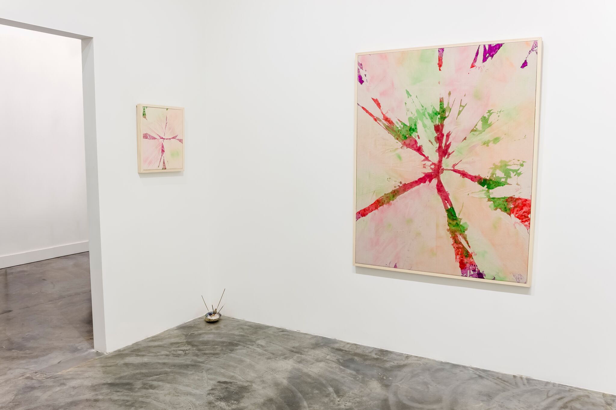 Contemporary gallery interior featuring abstract paintings with radiating splashes of red, pink, green, and purple watercolor on pale backgrounds, displayed on white walls alongside a sculptural object with protruding wire elements on the concrete floor.