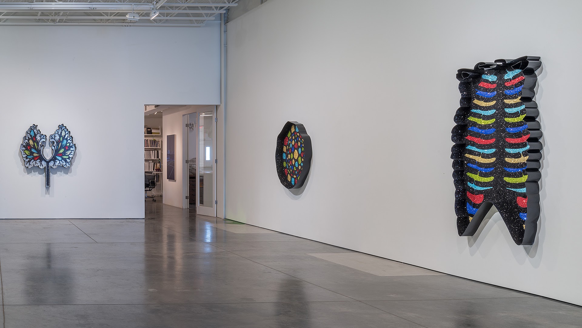 Contemporary gallery wall displaying three black sculptural forms embellished with multicolored glitter arranged horizontally. Left: symmetrical tree-like shape with blue and iridescent accents. Center: circular disc with densely packed colorful details. Right: tall vertical form resembling anatomical ribcage with layered bands of cyan, red, gold, and blue glitter against black textured surface. Polished concrete floor and white walls with industrial ceiling visible.