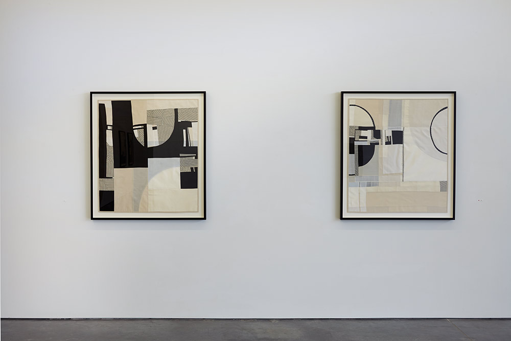 Two framed abstract textile artworks hang on a white gallery wall, each composed of layered black, white, and beige fabric pieces arranged in gestural, geometric compositions. The works feature translucent qualities created through fused fabric layers, with bold curved and angular forms emerging from striped and solid textile elements.