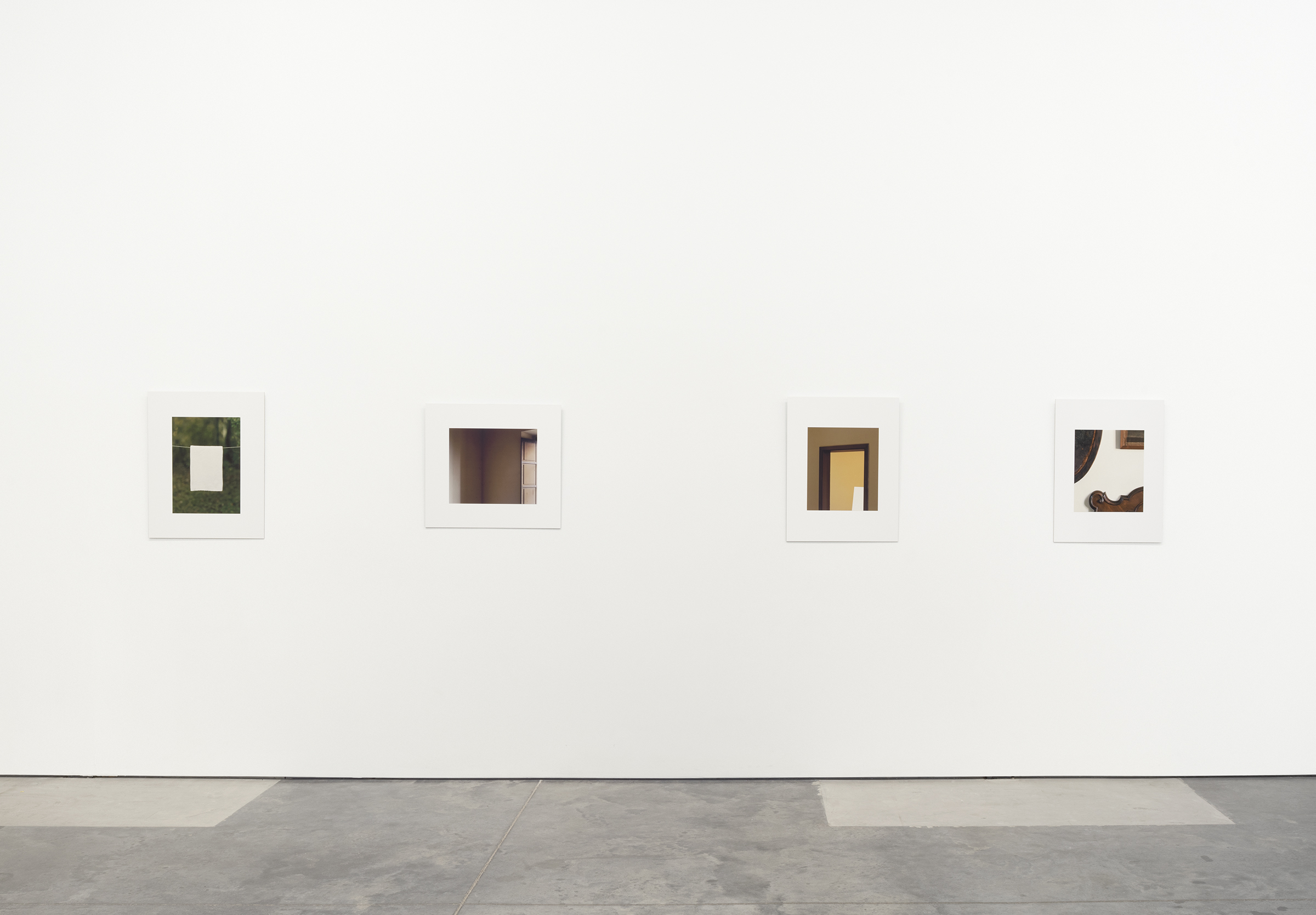 Four framed photographs mounted on a white gallery wall display geometric interventions within architectural and natural spaces. The images feature rectangular shapes of varying colors and materials—moss green, deep brown, ochre, and mixed tones—inserted into doorways, corners, and interior scenes, exploring the relationship between placement, perception, and constructed space.