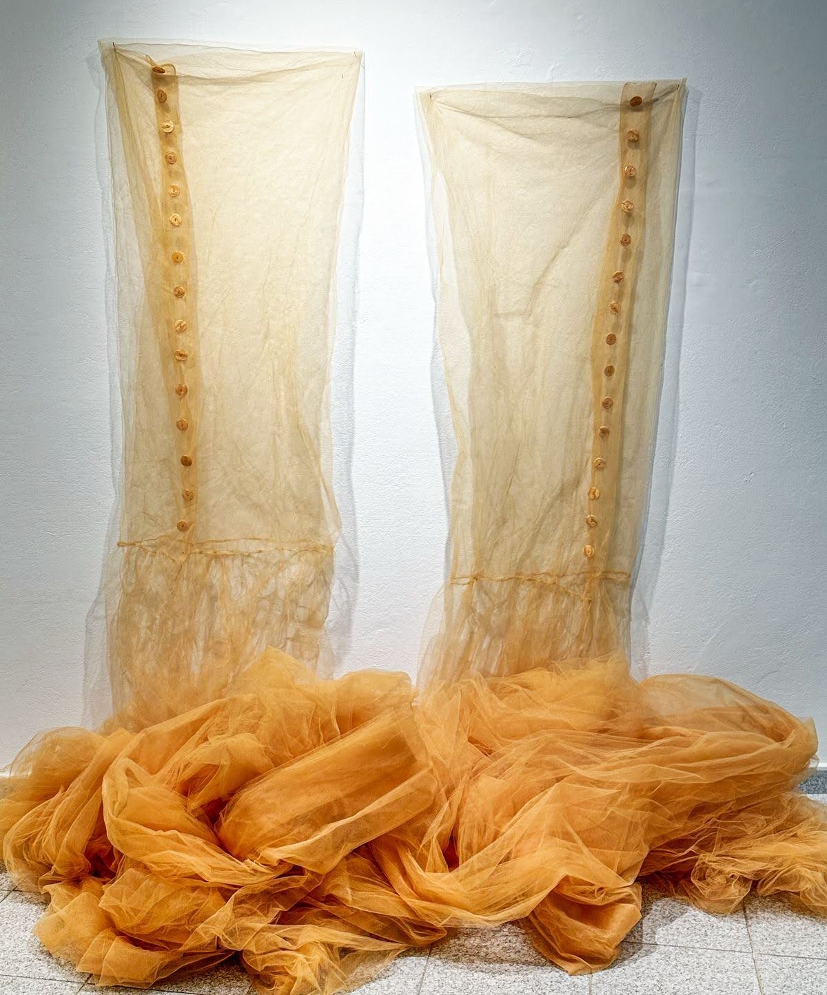 Two translucent cream-colored tulle panels hang vertically against a white wall, each fastened with a vertical line of golden buttons, while voluminous folds of golden-orange tulle pool and drape across the floor below, creating a composition that explores the tension between constraint and liberation through layered, sheer fabric.