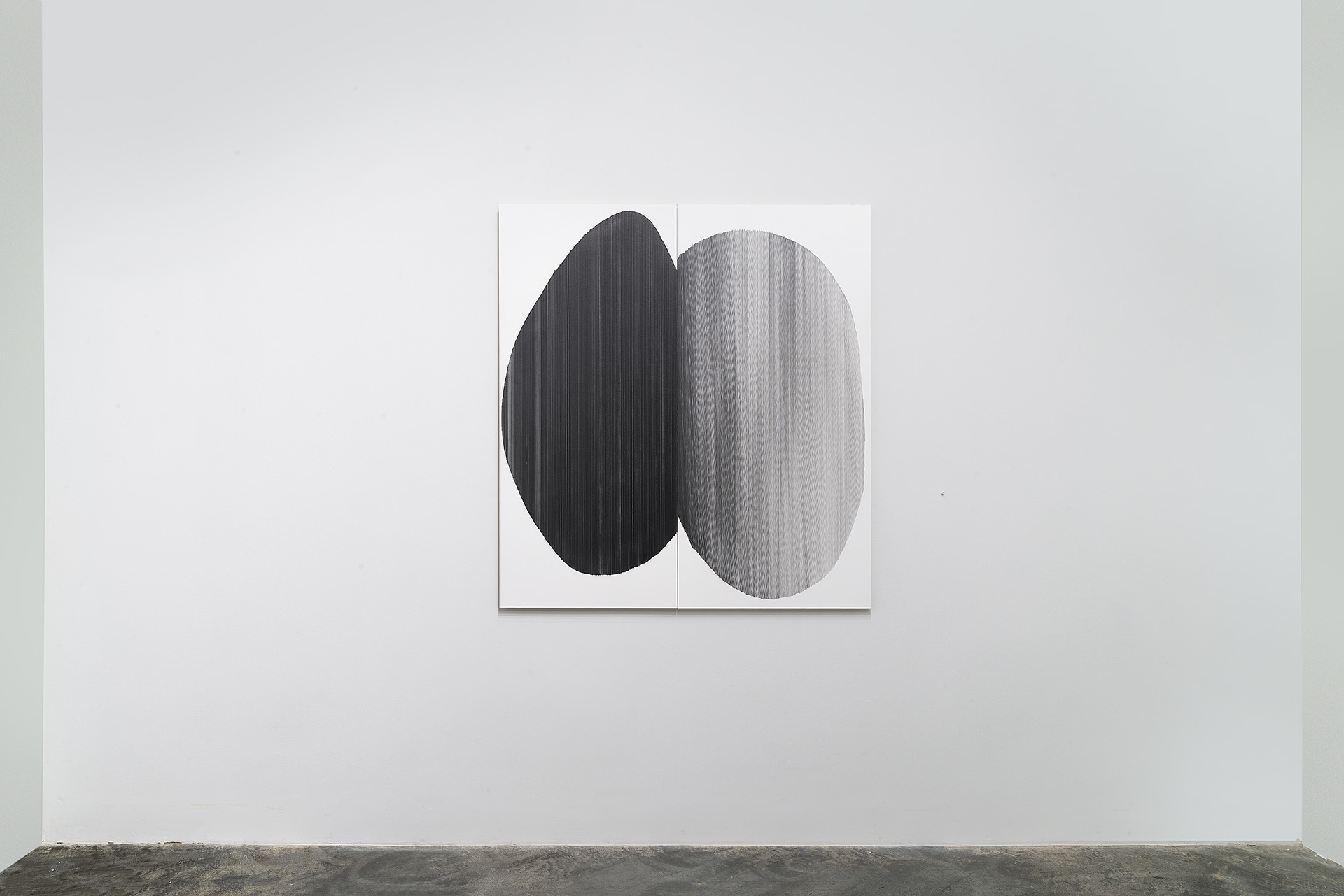 Two large oval forms, one dark charcoal and one light gray, face each other on a white two-panel surface. Both ovals feature subtle linear surface textures and are rendered in hand-drawn graphite, creating a sense of weight and dimensionality against the gallery wall.