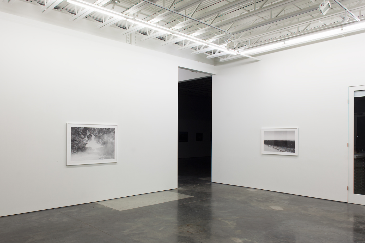 A minimalist gallery interior with white walls and polished concrete floor displays two black-and-white framed photographs: one depicting a misty wooded landscape on the left wall, and another showing a vast, flat field on the right. A large black doorway opens in the center wall, and industrial ceiling fixtures with exposed beams illuminate the sparse exhibition space.