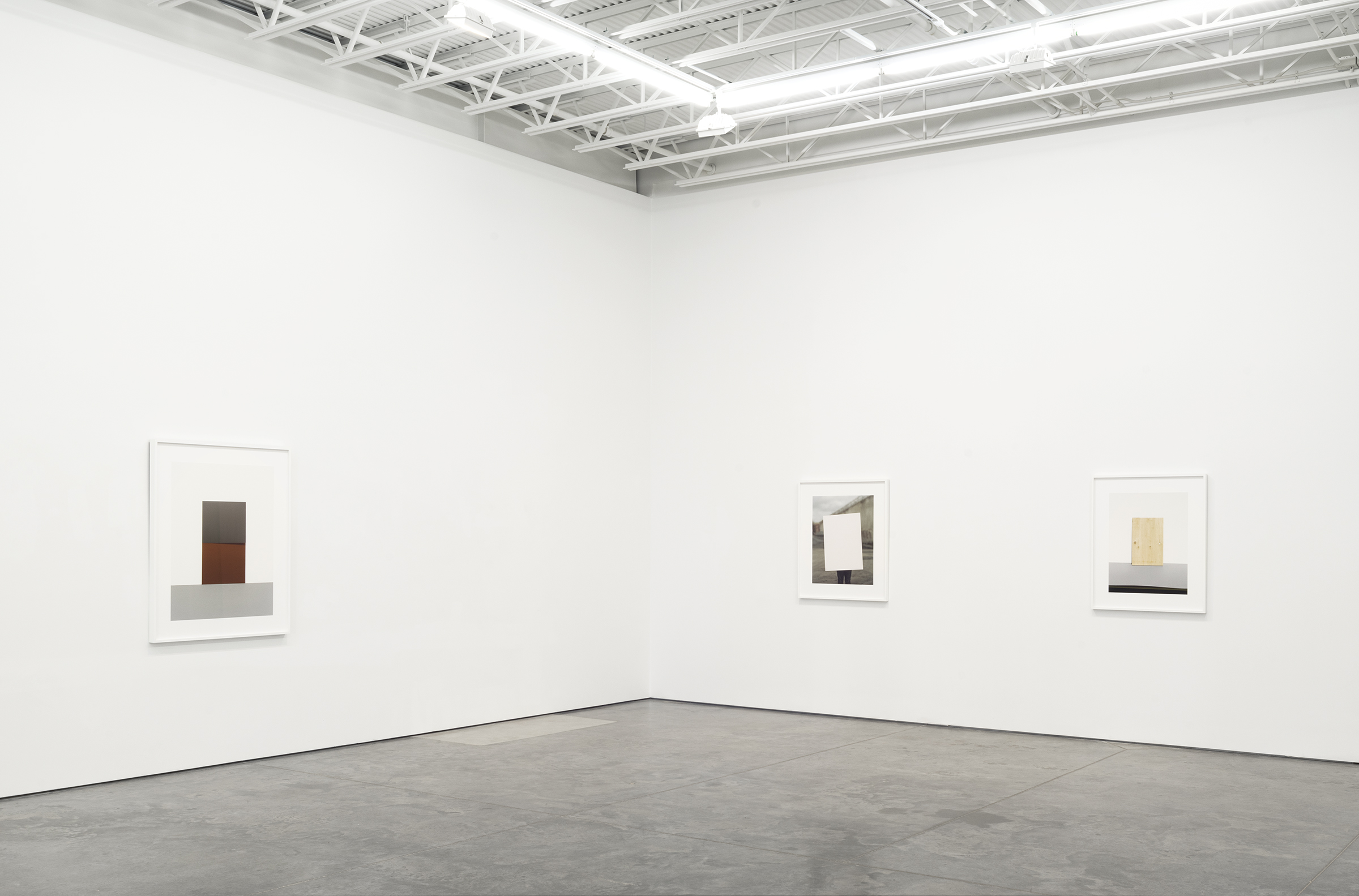 Three framed photographs hang on a white gallery wall, each featuring geometric compositions of colored rectangles and planes arranged within minimalist settings. The leftmost image shows stacked brown and gray forms, the center depicts a light rectangular aperture, and the right presents layered tan, gray, and black elements. The works explore abstract spatial relationships and the interplay between constructed and observed forms.