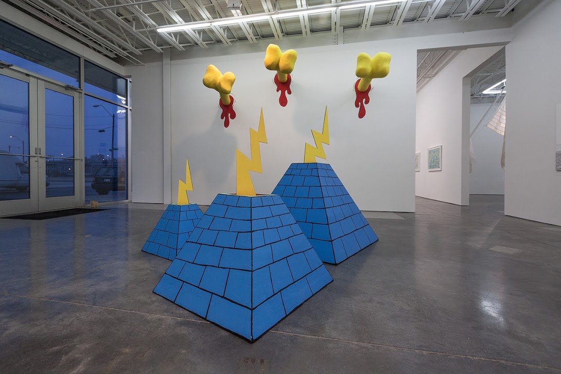 Three blue pyramids with yellow lightning bolts and circular protrusions sit on a polished concrete floor in a minimalist gallery space, while three yellow organic forms with red dripping elements hang on the white wall above, evoking playful yet unsettling imagery reminiscent of 1970s cartoon aesthetics.